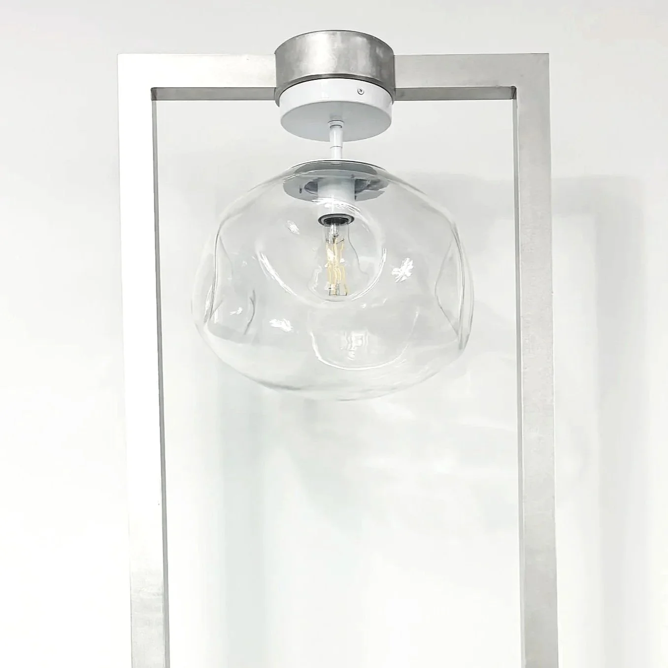 Modern ceiling light fixture with a clear, irregularly-shaped glass shade and a visible filament bulb, mounted on a minimalistic metal frame.