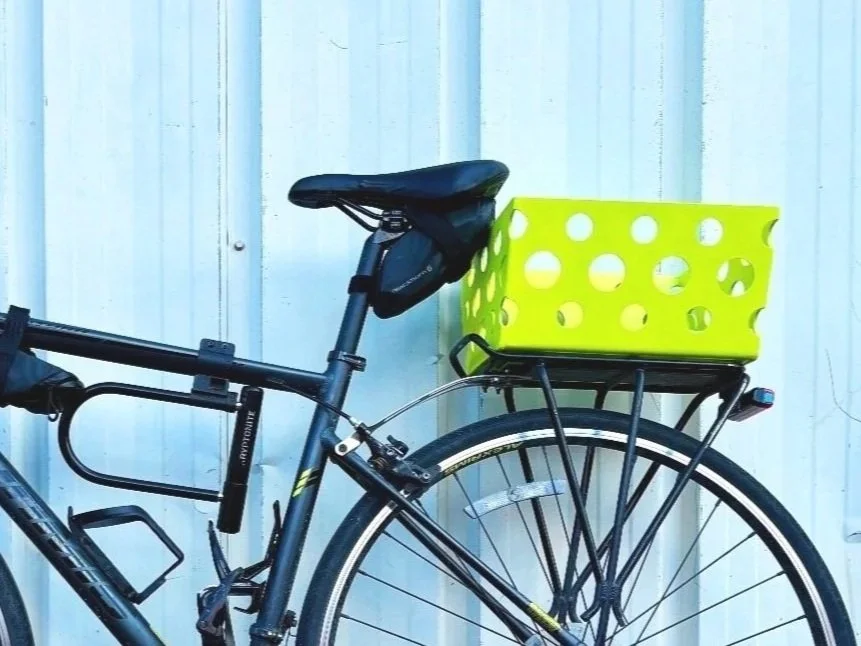 A black bicycle with a yellow basket attached to the rear, standing against a white corrugated metal wall.