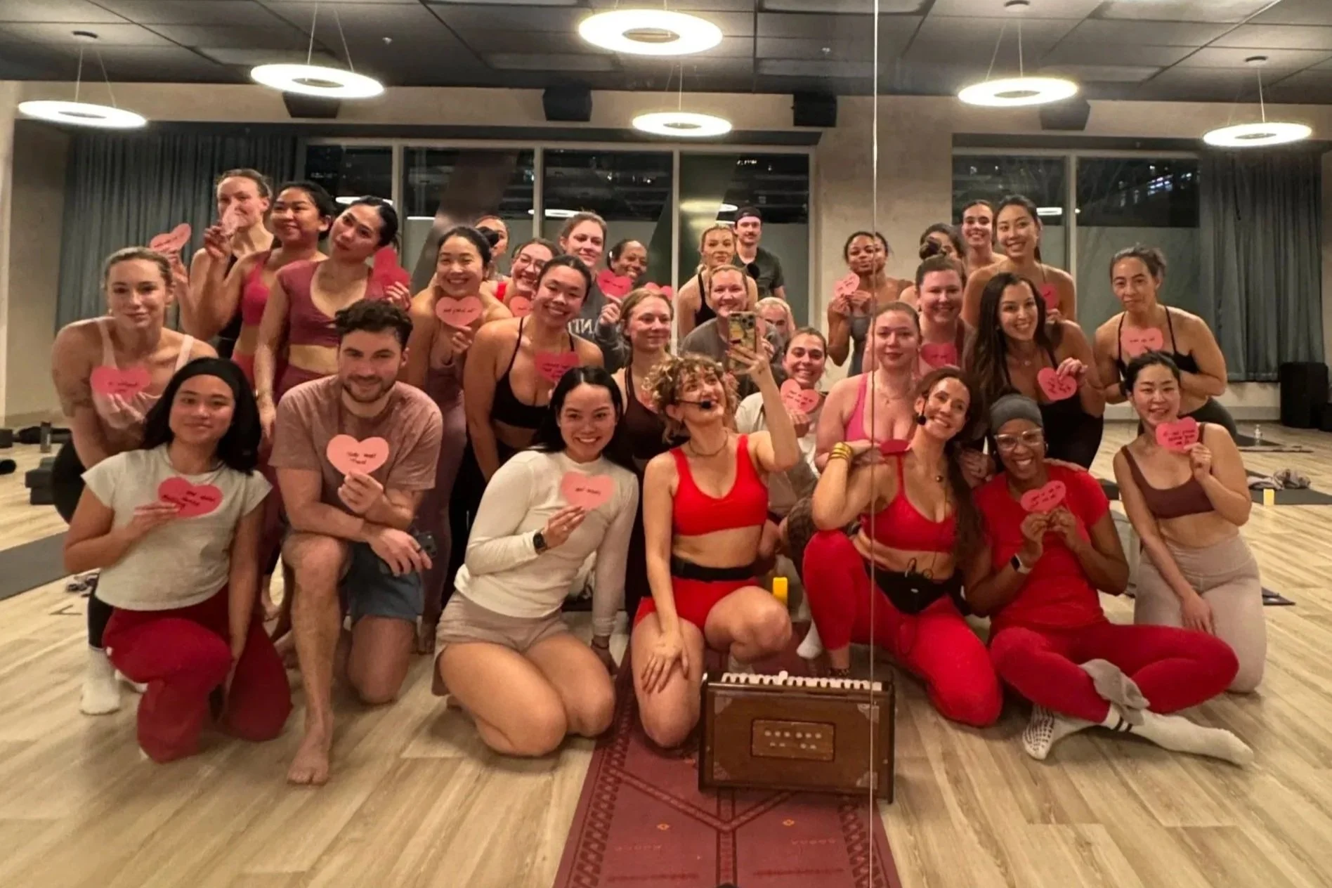 A group of woman and men in a dance or fitness class holding pink paper hearts, some with handwritten notes, posing in a studio with a wooden floor and modern lighting.