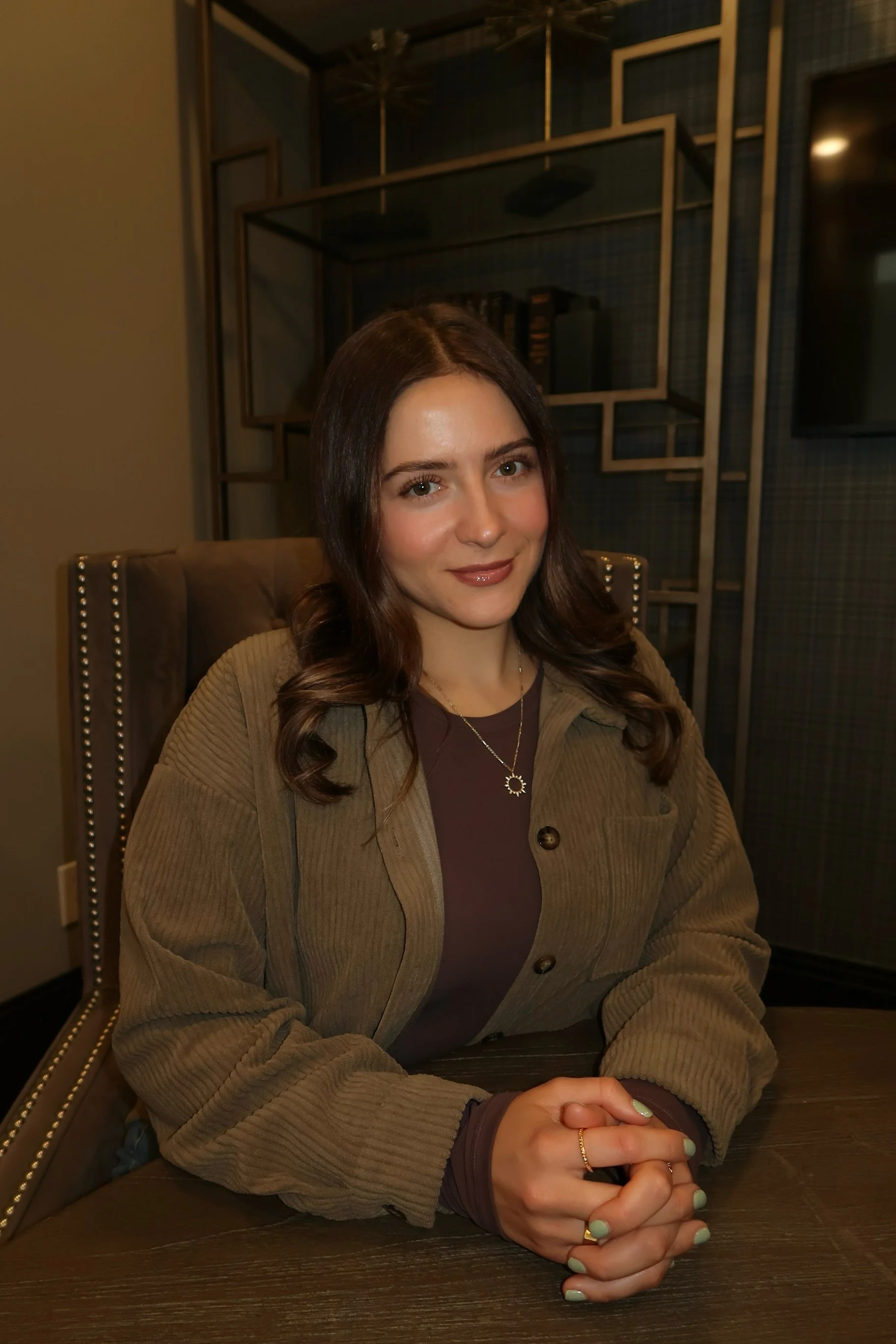 Young woman with brown hair sitting at a table in a modern interior, wearing a brown jacket and a necklace, smiling at the camera.