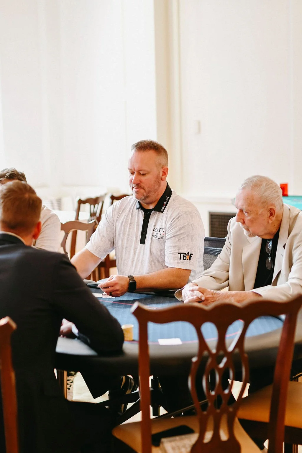 Men playing poker at a card table in a bright room, with focus on a man wearing a white shirt with logos.