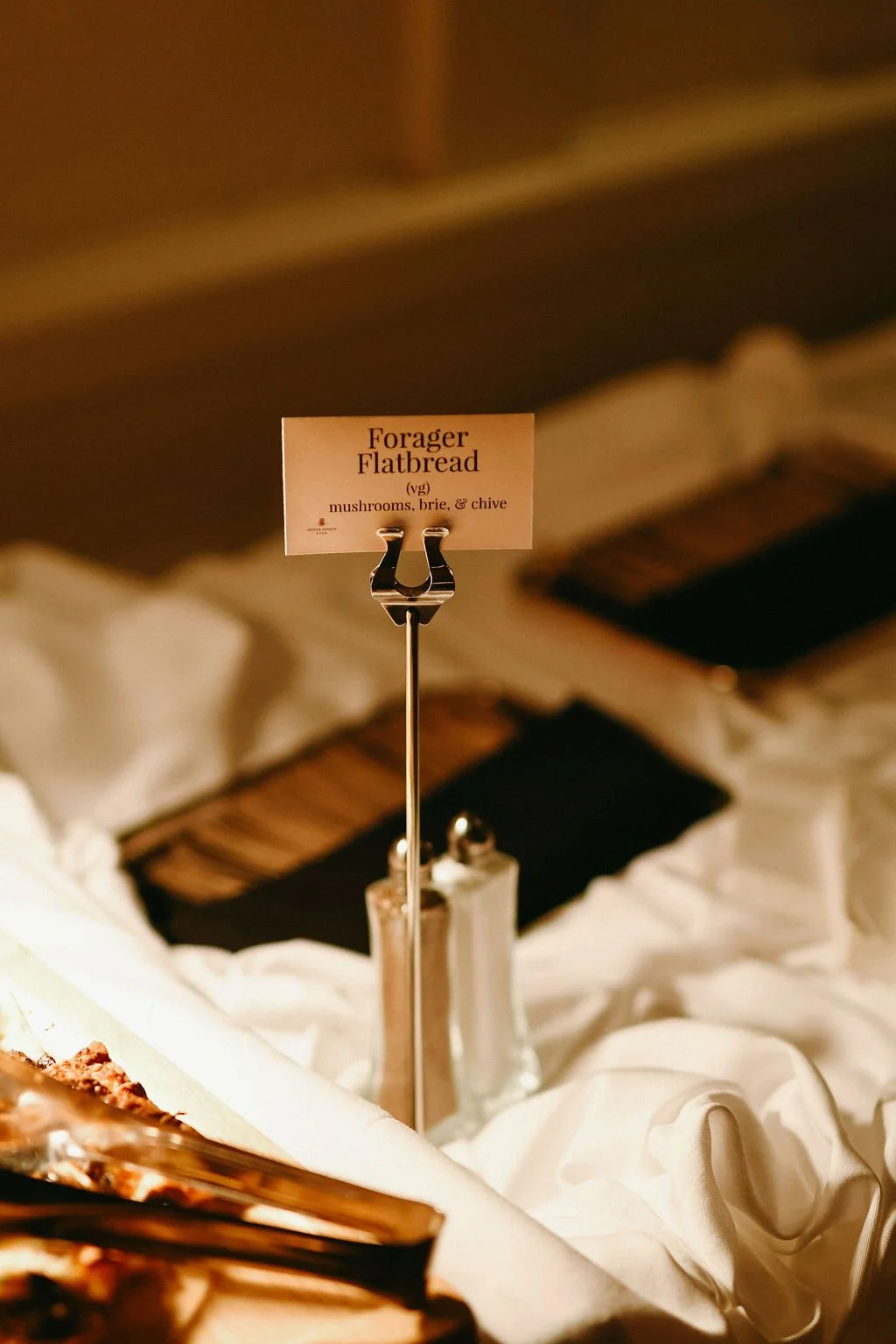 A sign displays 'Forager Flatbread' with ingredients mushrooms, brie, and chive, placed on a metal stand over a white cloth at a buffet or food display.