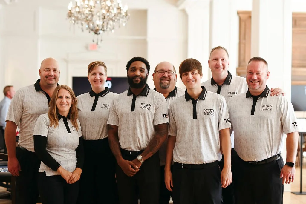 Group of seven people, six men and one woman, wearing matching gray and black shirts with 'POKER PURPOSE' logo, standing together indoors with elegant decor and chandelier in the background.