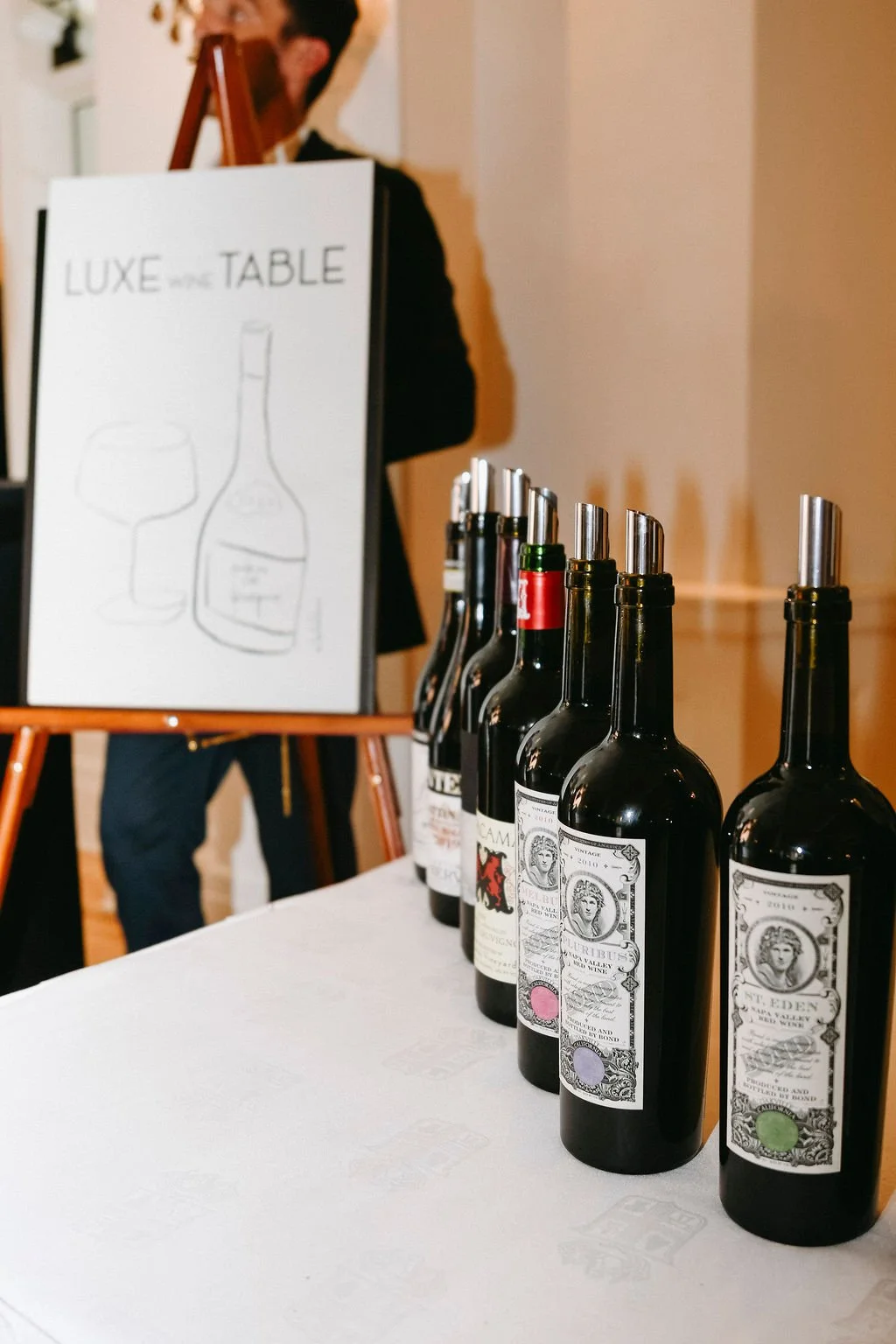 Line of wine bottles with metal pour spouts on a table, with a sign that reads 'LUXE the TABLE' and a person standing in the background.