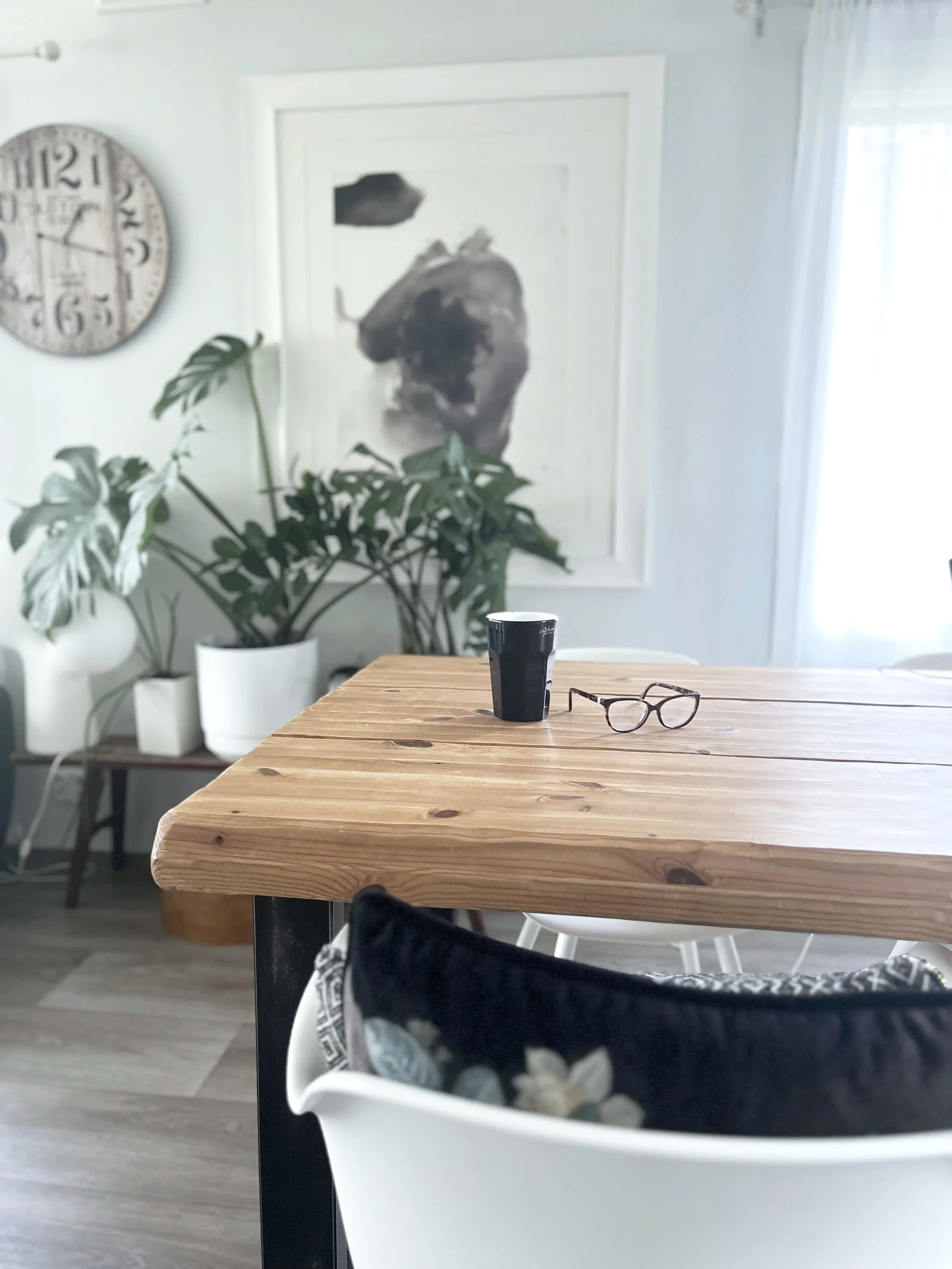 Kuvaus keittiö- tai ruokailutilasta, jossa on puinen pöytä. Pöydällä on muovimuki ja silmälasit. Taustalla on iso monstera kasvi ja abstrakti taideteos seinällä. Myös kello on seinällä.