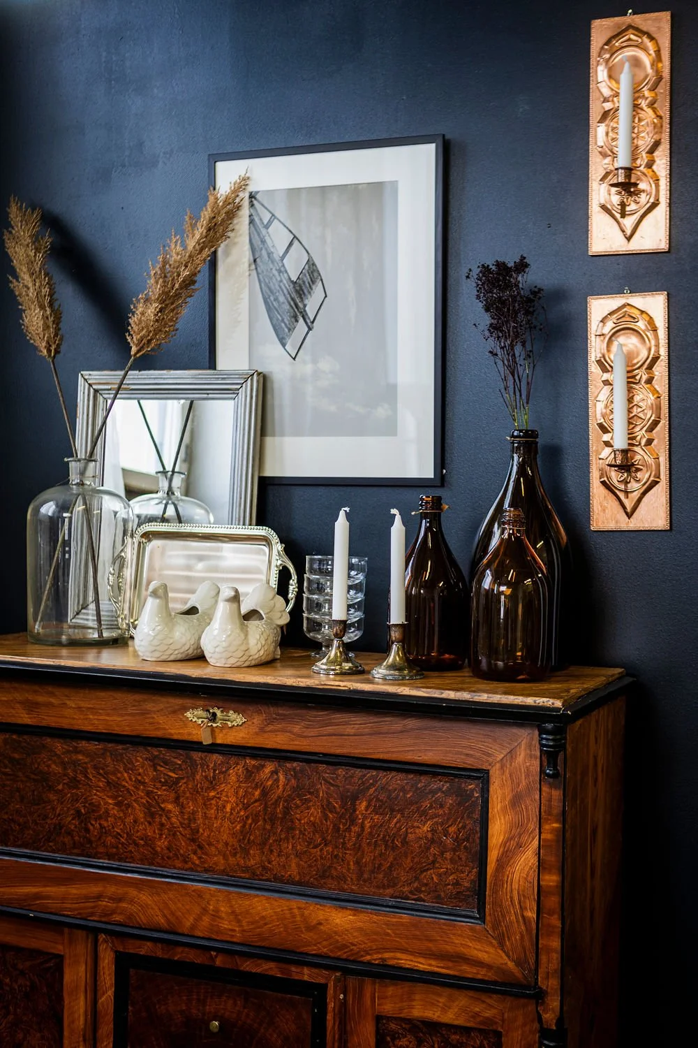 Kuvassa on tummansinisellä seinällä oleva kuva, jalallinen kynttilä ja koriste-esineitä, kuten vaaleita pöllölasiesineitä, maljakoita ja kynttiläjaloja, sekä peili ja taideteoksia kynttilöillä.