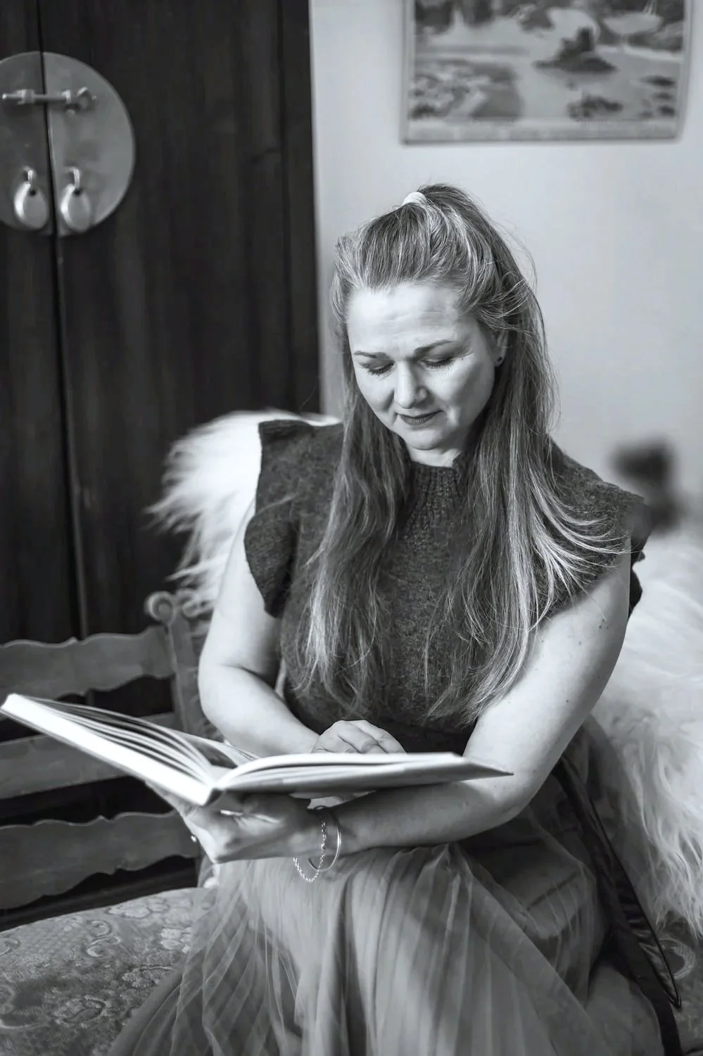 Nainen lukee kirjaa sisätiloissa, takana puinen ovea ja taulu seinällä.