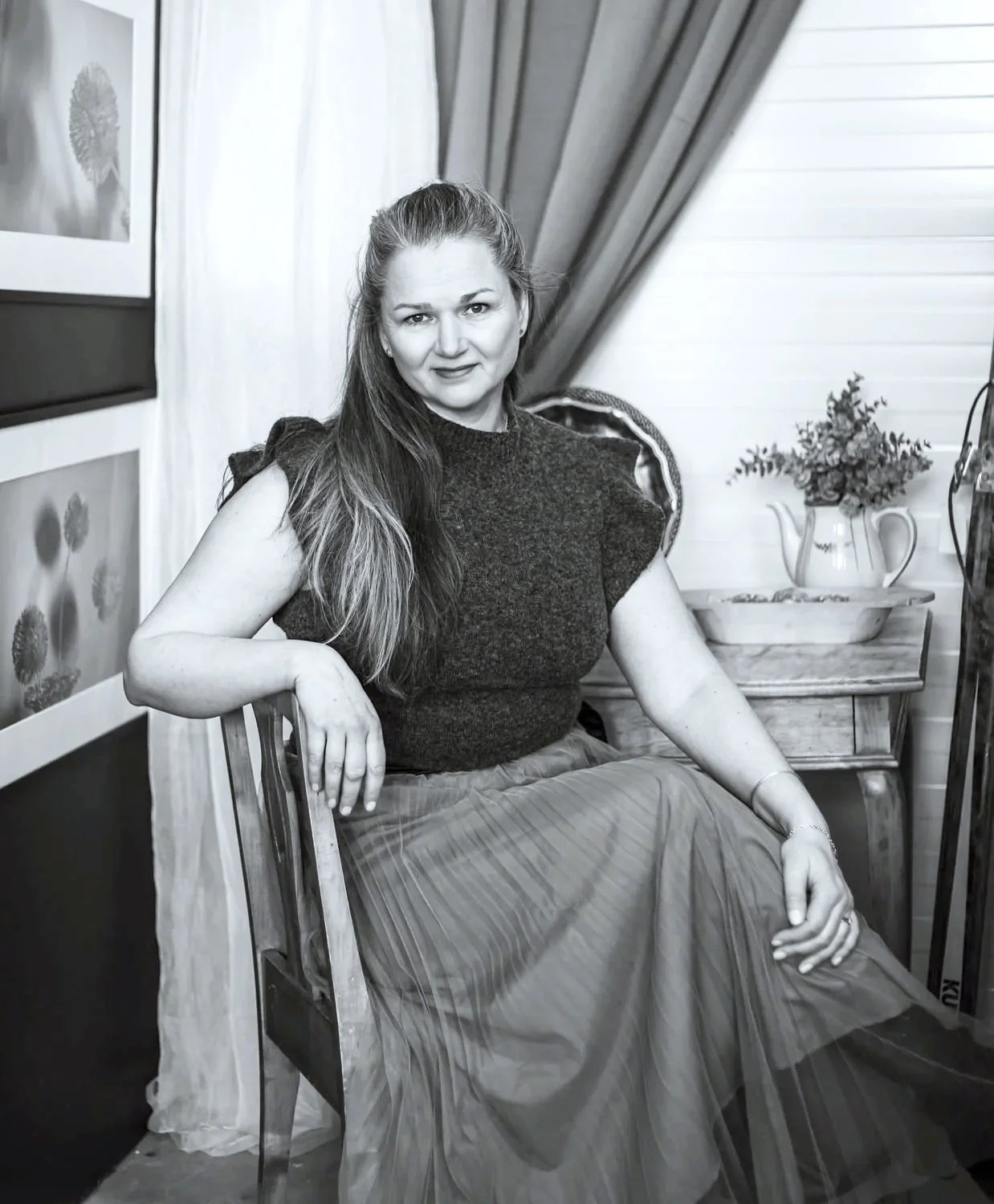 Mustavalkoinen valokuva naisesta istumassa tuolilla, taustalla kasveja ja verho, hymyilevä nainen pitkästä hiuksista.