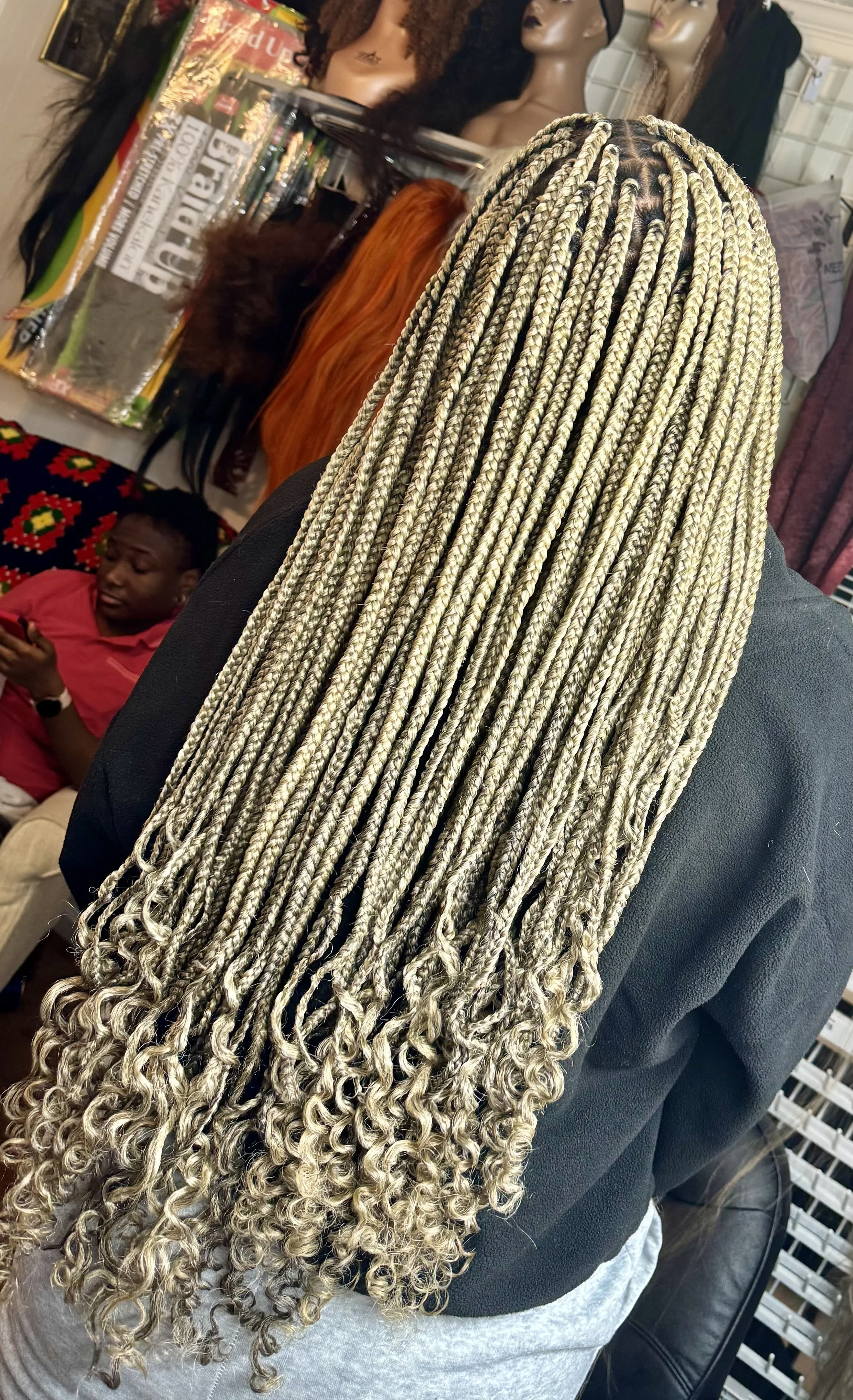 Person with long, light-colored braided hair in a salon, with curly tips, surrounded by hair products and mannequins.