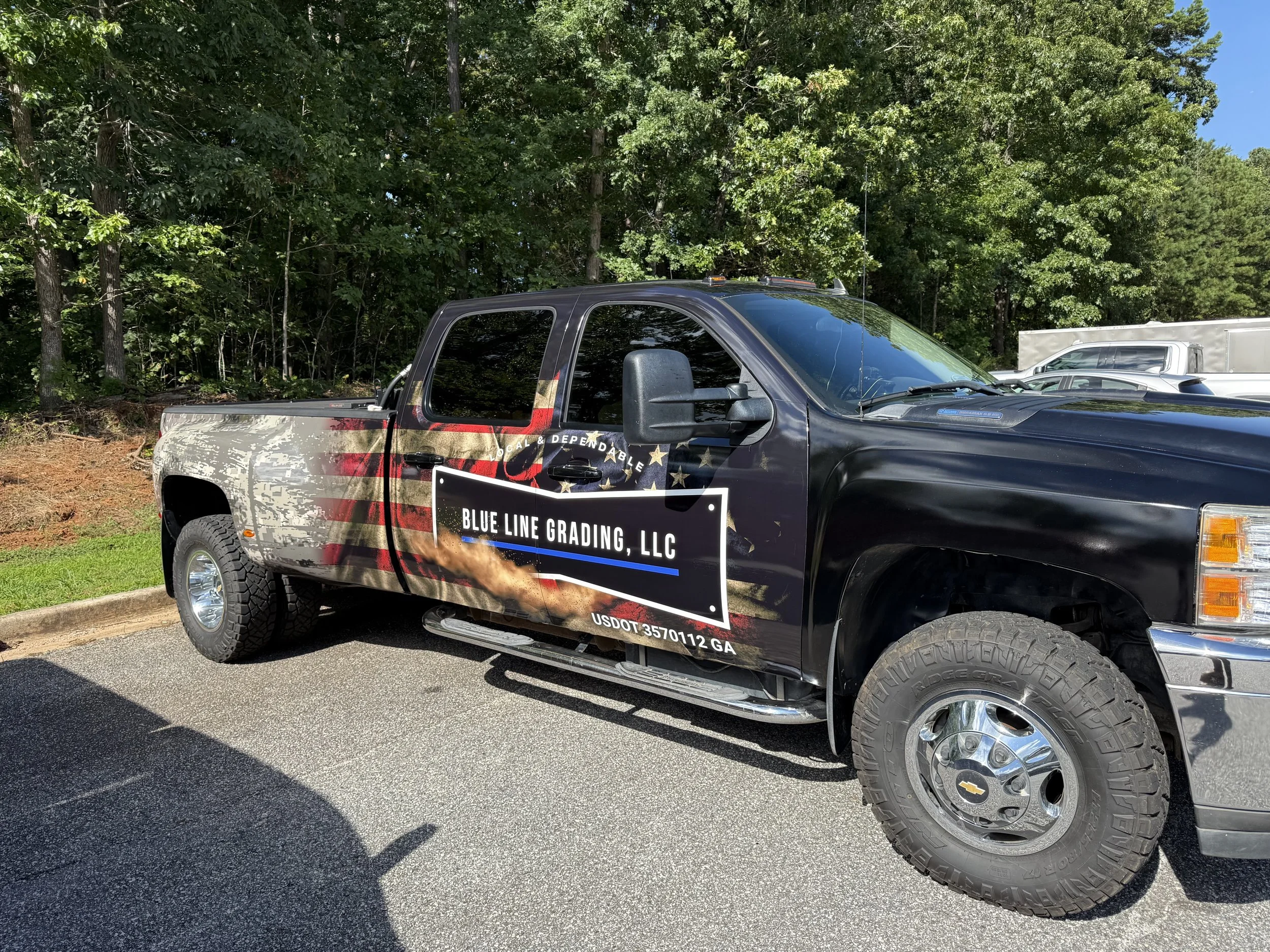 Black Chevrolet pickup truck with a commercial graphic for Blue Line Grading LLC parked outdoors near a wooded area.