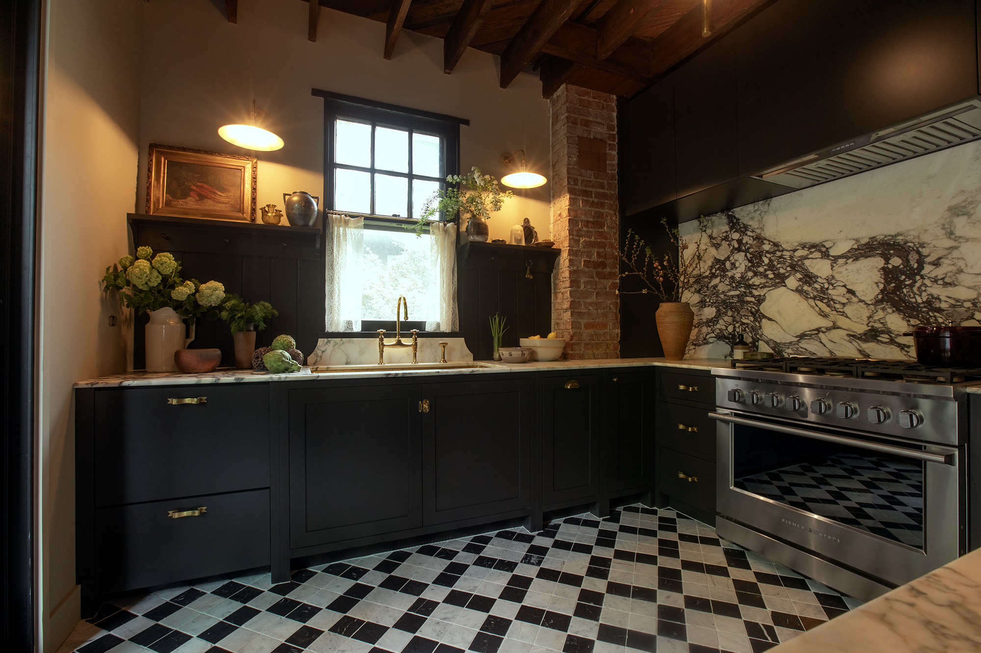 A modern kitchen with black cabinets, brass handles, and a marble backsplash. There is a window above the sink with a curtain, and various decorative items including vases and plants. A stainless steel oven is on the right, and the floor has black an