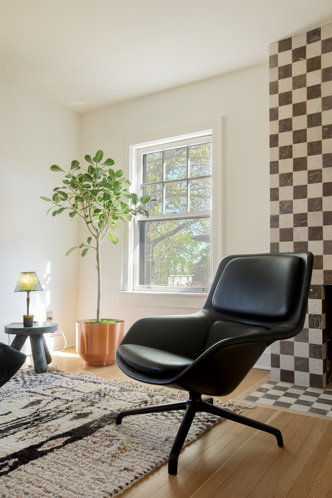 Living room with a black modern armchair, a large potted plant near a window, a small side table with a lamp, a patterned rug, and a wall with a checkered brick design.