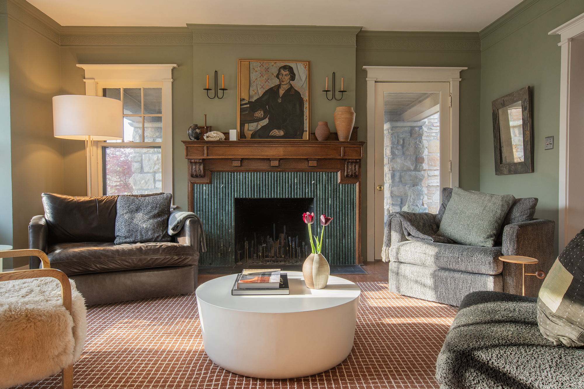 Living room with a stone fireplace, a brown leather sofa, a gray fabric sofa, a white round coffee table with a vase of pink tulips, a large floor lamp, green walls, and artwork above the fireplace.