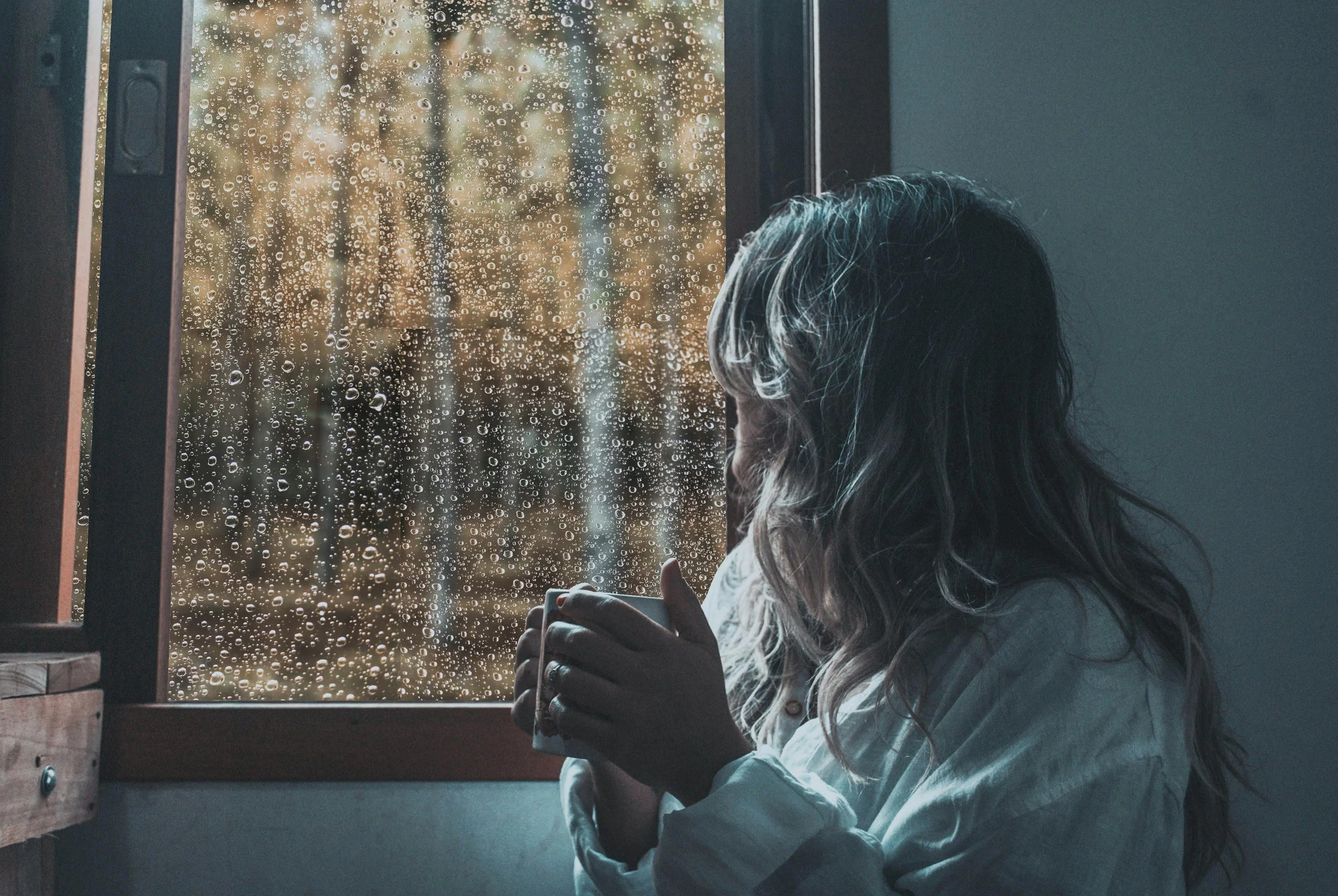 Woman sitting alone looking out a rain-covered window holding a mug, illustrating the emotional disconnection and isolation common in complex PTSD