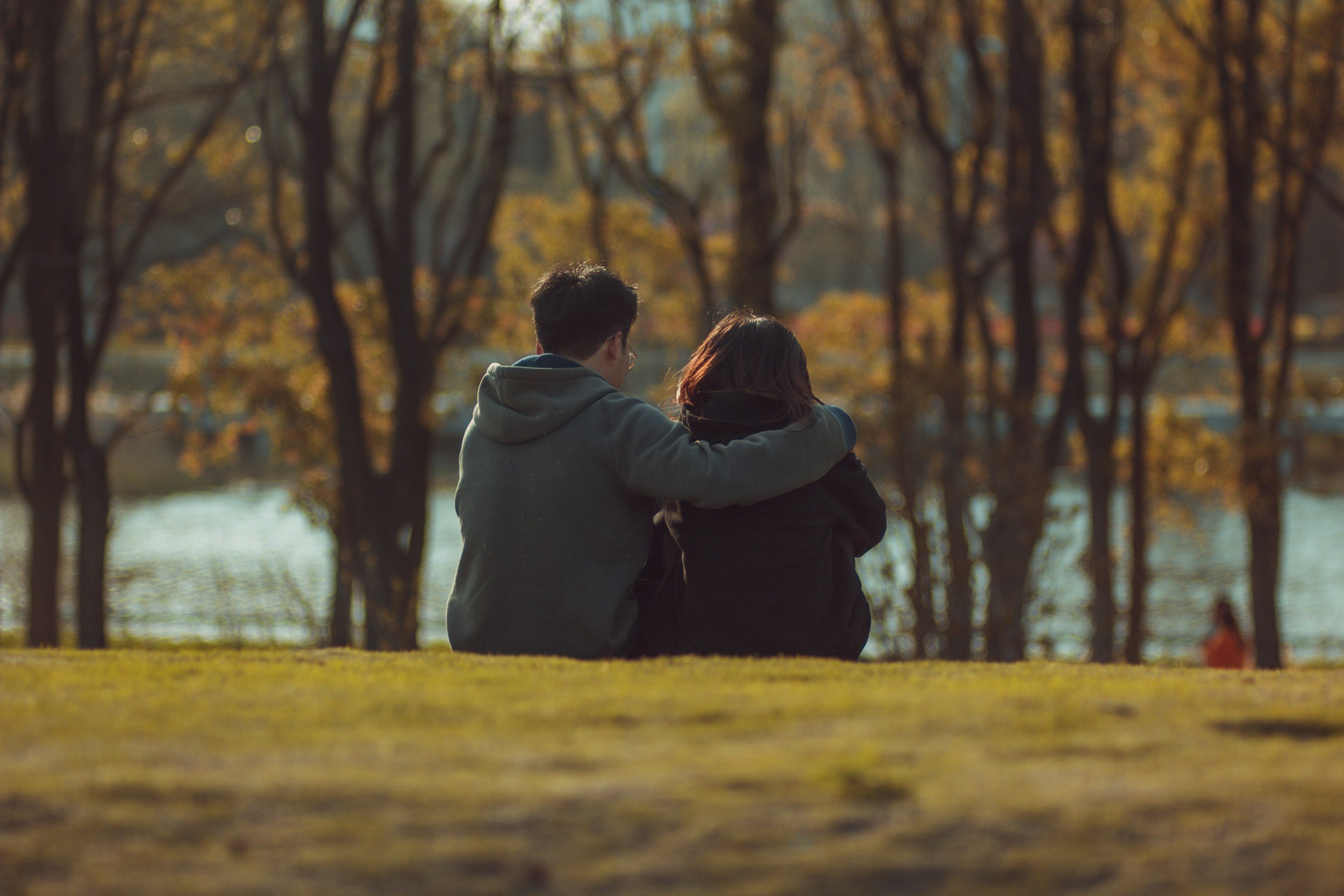 A young man and woman sitting close together by a lake, with the man having his arm around the woman's shoulder, in an autumn park surrounded by trees with fall foliage.