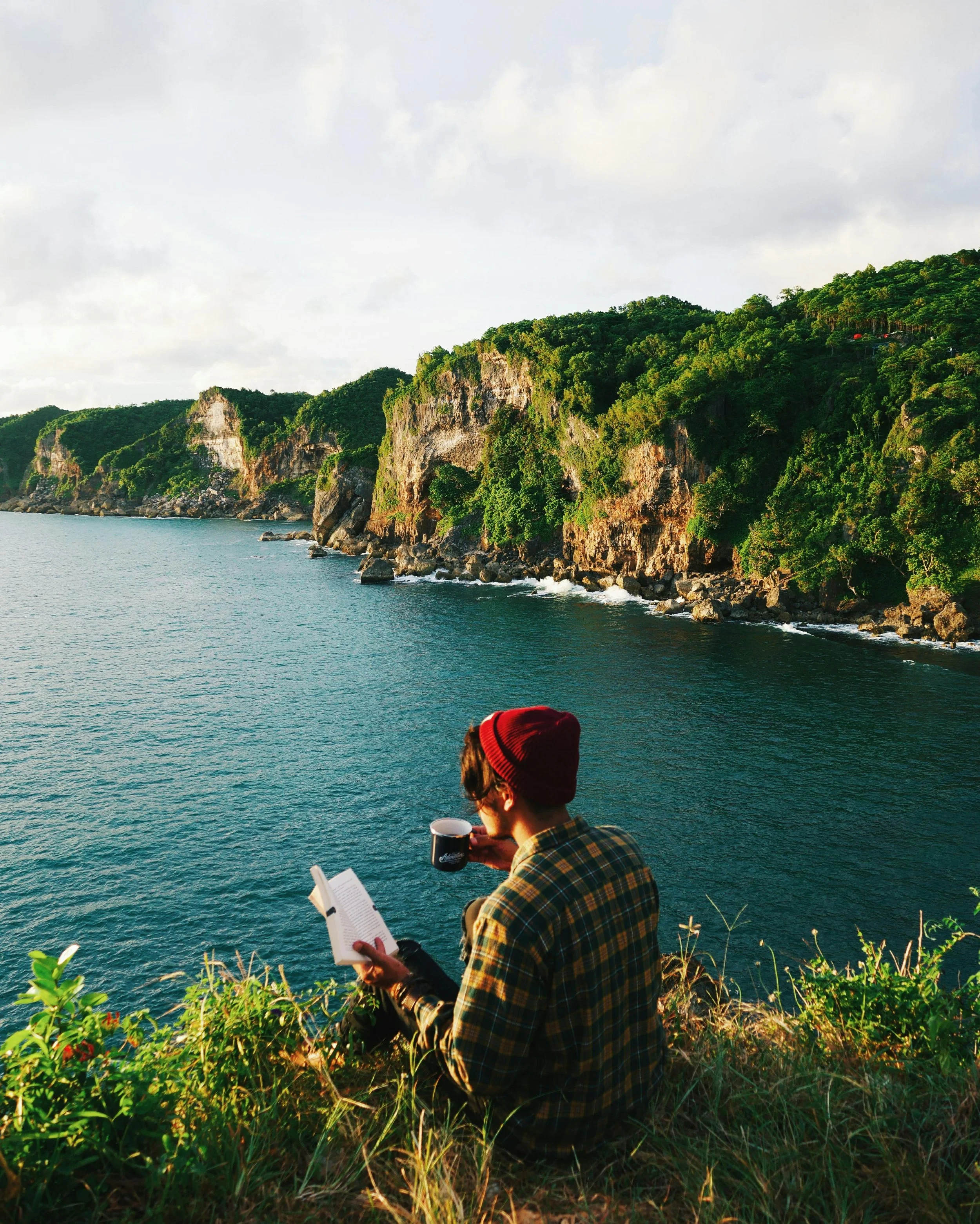A person wearing a red beanie and glasses, dressed in a yellow plaid shirt, sitting on the grassy verge of a cliff, reading a book and holding a cup of coffee, with the ocean and green cliffs in the background during the day.