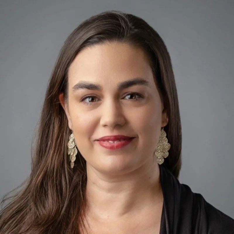 Portrait of a woman with long brown hair, wearing earrings and a black top, against a gray background.
