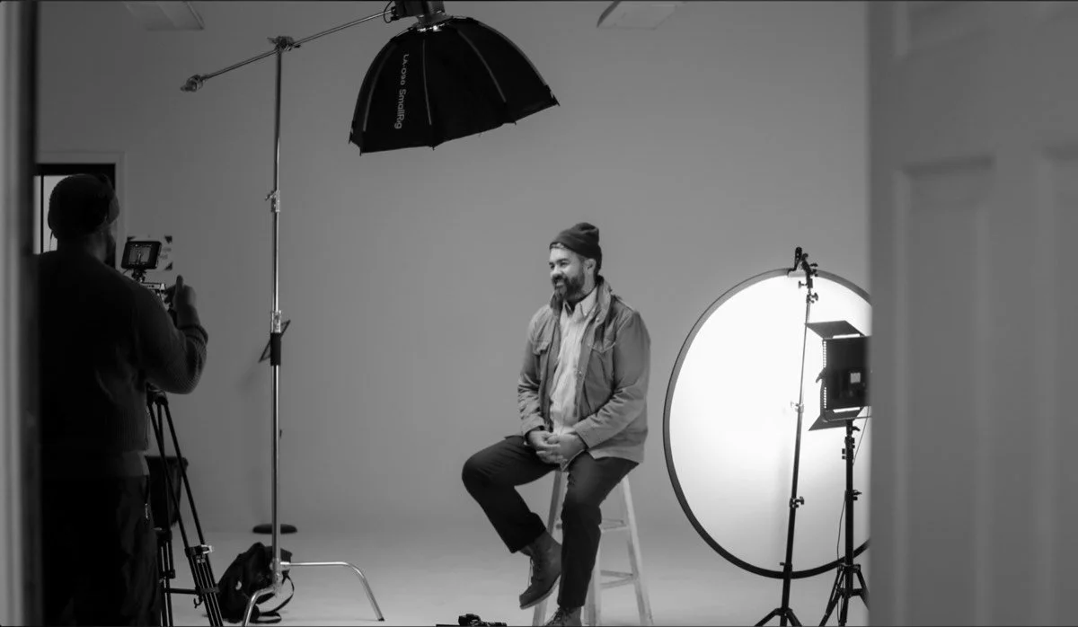 Behind the scenes of a KLTRD MEDIA studio interview: camera operator filming a seated subject with softbox and reflector lighting