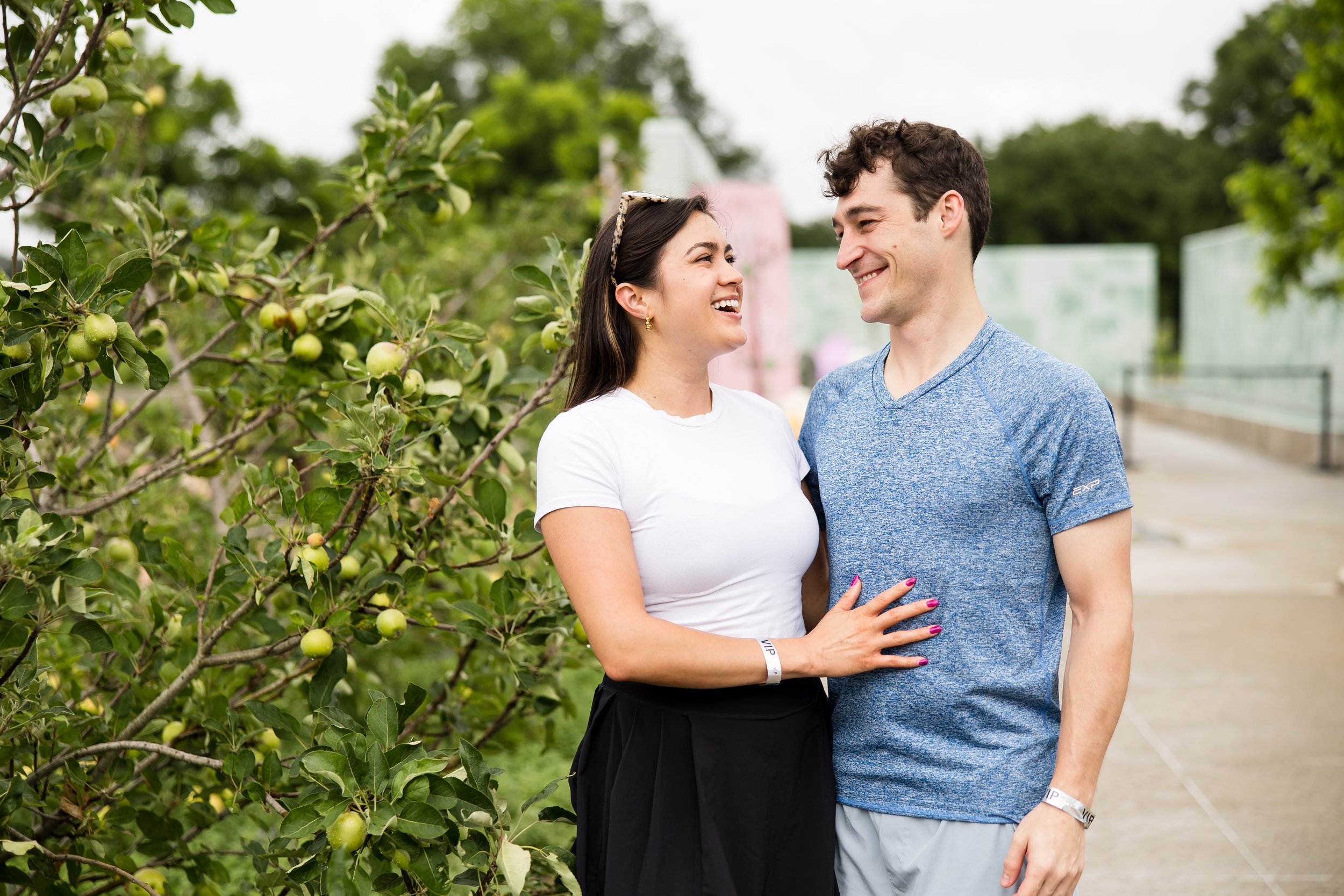 A couple standing near an apple tree, smiling at each other. The woman is wearing a white shirt and black skirt, while the man is in a blue T-shirt. The background features greenery and a walkway.