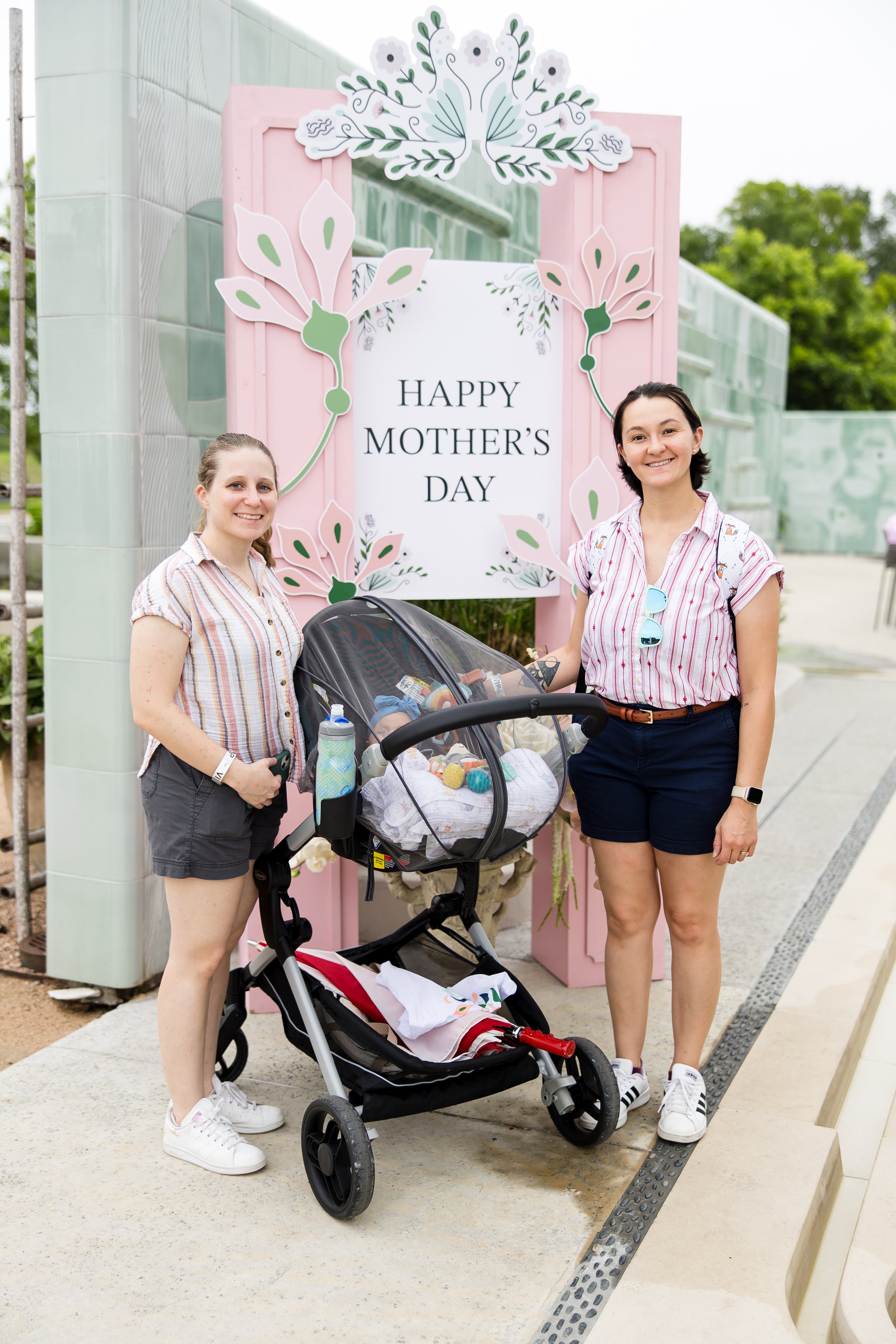 Two people standing beside a decorated sign reading "Happy Mother's Day" with a stroller containing a baby. Both are wearing casual attire and smiling.