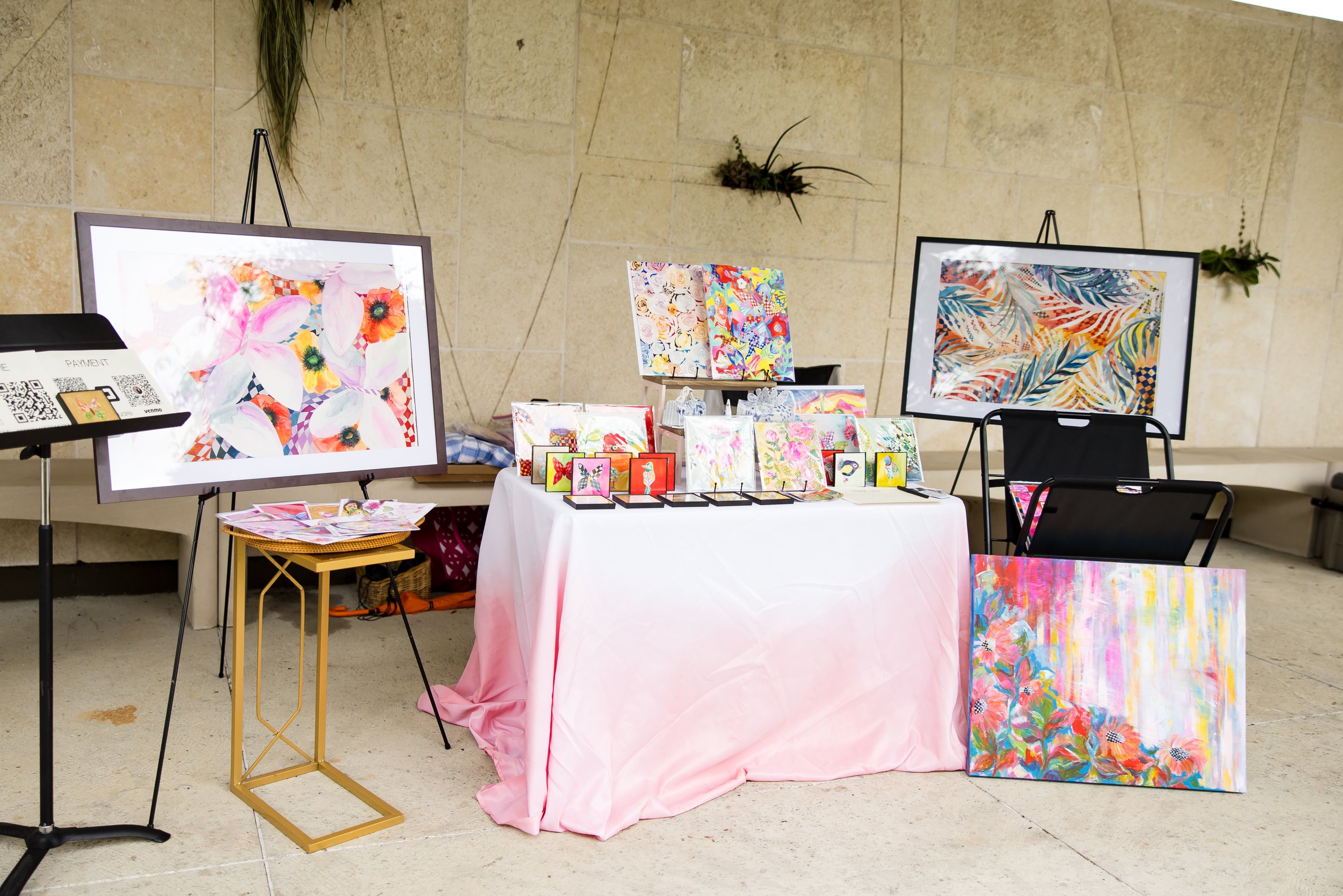 Art display with colorful paintings, framed floral and abstract images, and small artworks on a table with a pink tablecloth at a market or exhibit.