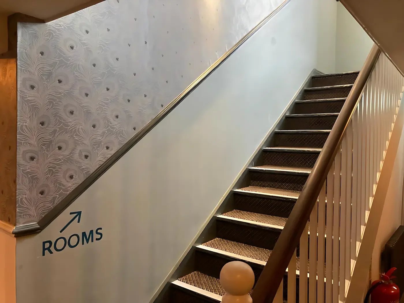 Indoor staircase with carpeted steps, wooden handrail, white spindles, and a wall with peacock feather wallpaper. An arrow pointing to the left indicates the direction to the rooms.
