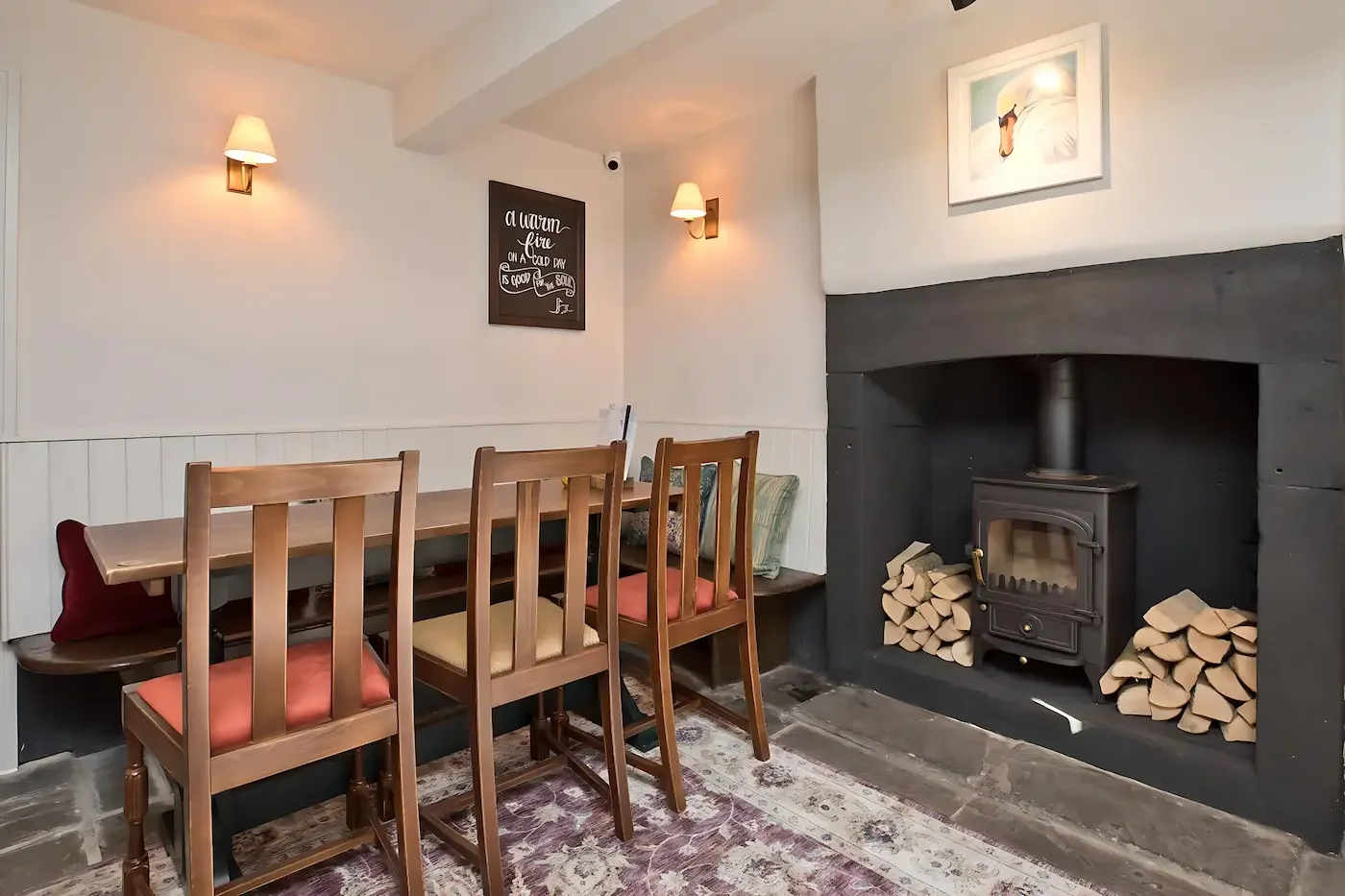 A cozy dining nook with a wooden table and four chairs, a fireplace with wood stored on both sides, and decorative wall art and lights.