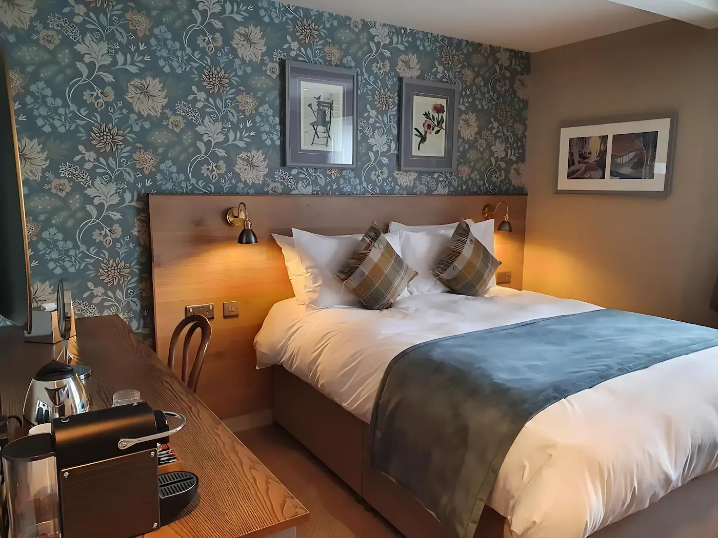 A hotel room with a neatly made bed with white sheets, two pillows, and a blue bed runner. The headboard is light wood, flanked by two wall-mounted reading lamps. The wall behind the bed has blue floral wallpaper, decorated with three framed pictures. To the left is a desk with a kettle, cups, and a mirror.