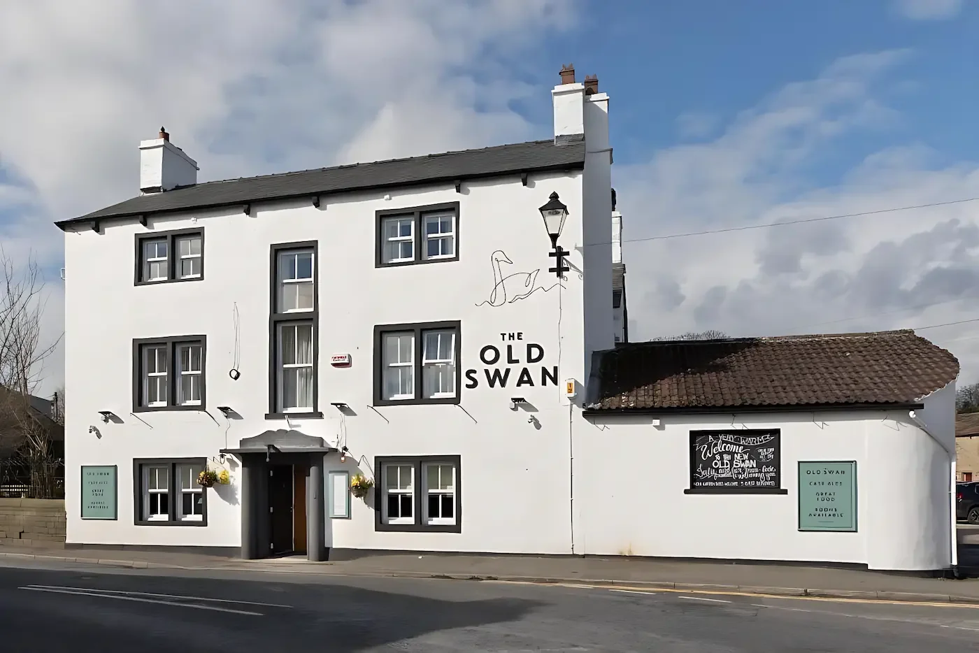 A white multi-story building with black window frames and a black door, named 'The Old Swan' pub, with hanging flower baskets and a mural of a swan on the exterior wall, located on a street with a cloudy sky overhead.