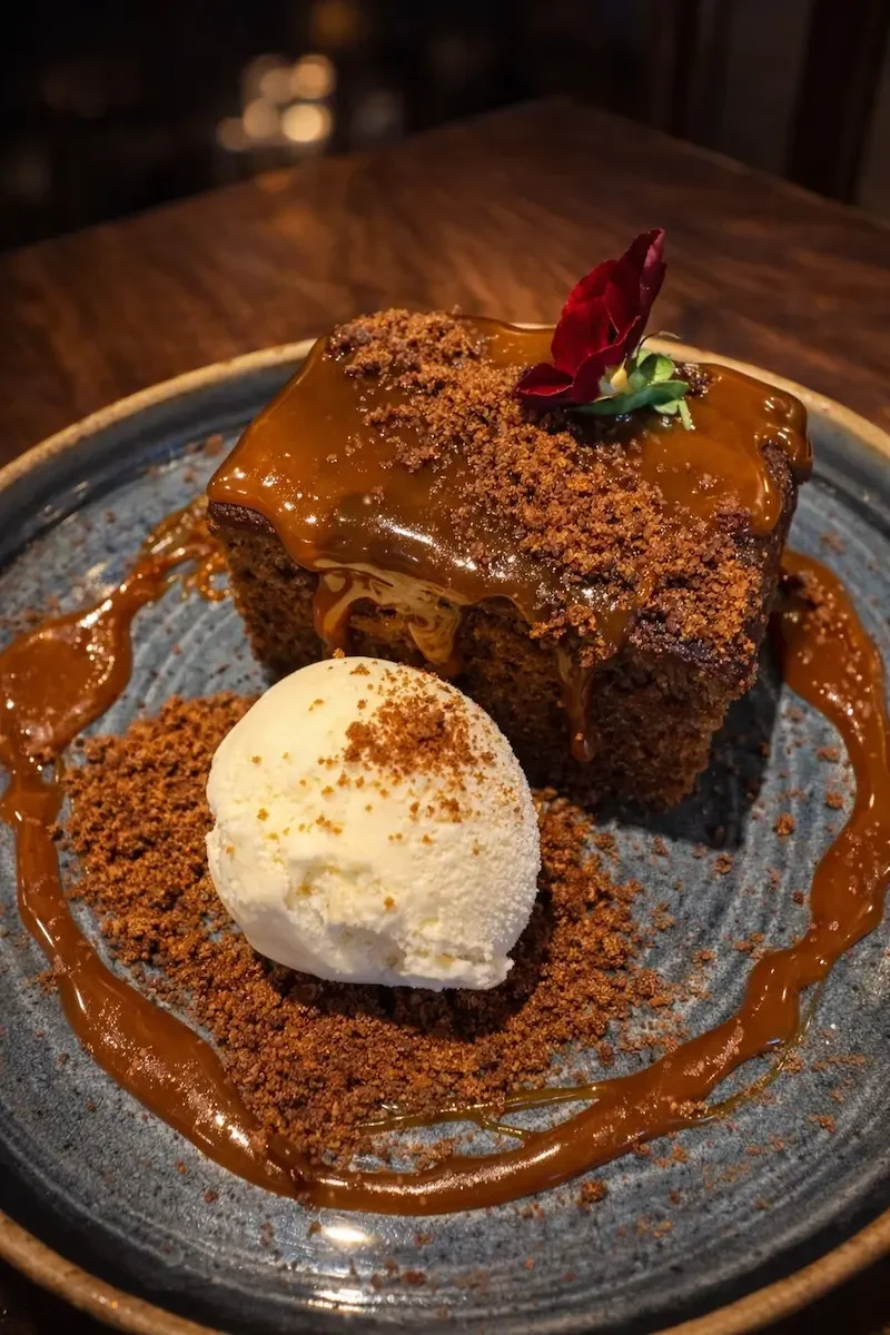 A slice of chocolate cake with caramel glaze, garnished with a red edible flower, served with a scoop of vanilla ice cream and caramel sauce on a blue plate.