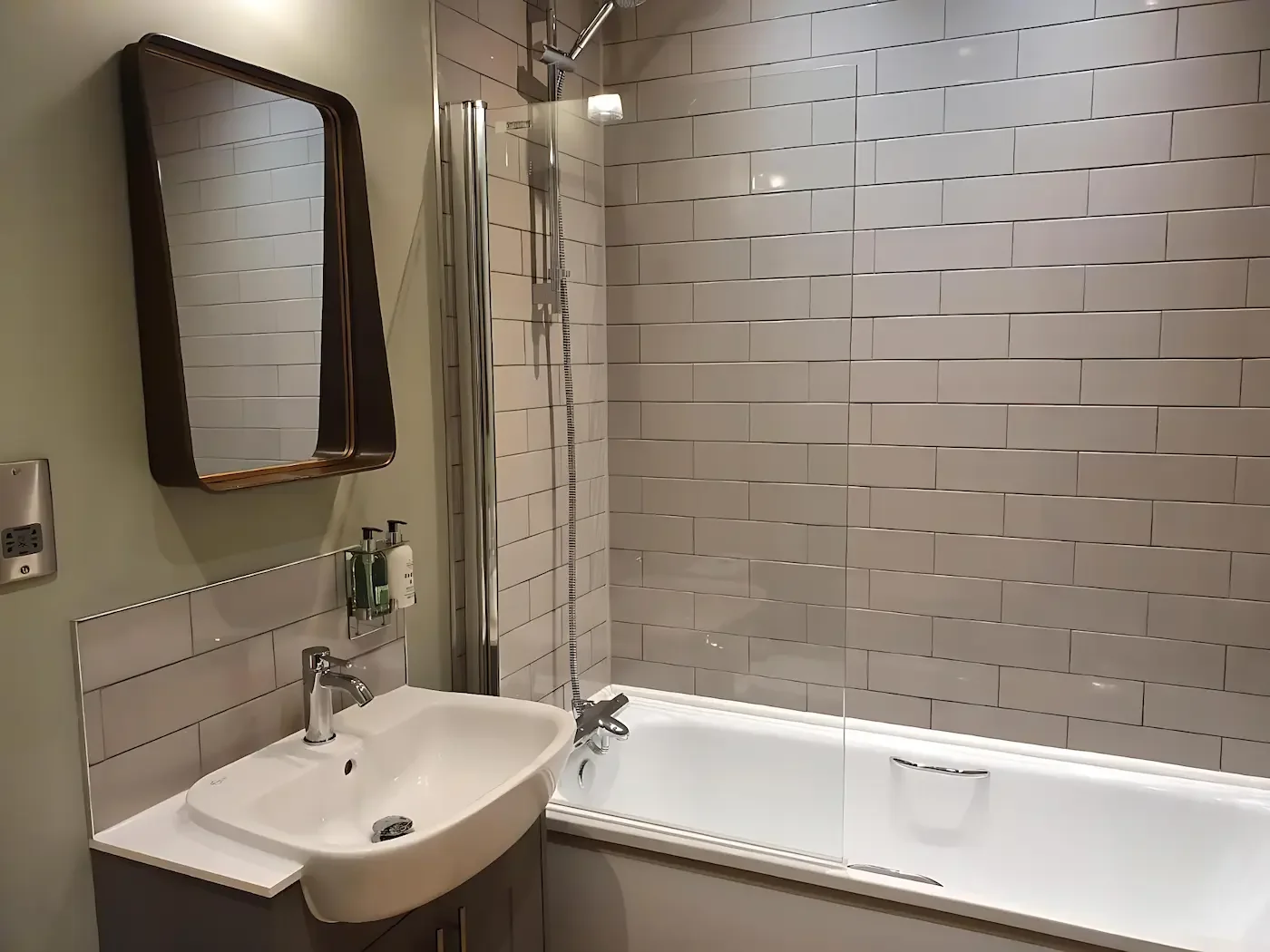 Modern bathroom with a sink, mirror, and a bathtub with a glass shower door. The wall features beige tiles and a soap dispenser is mounted above the sink.