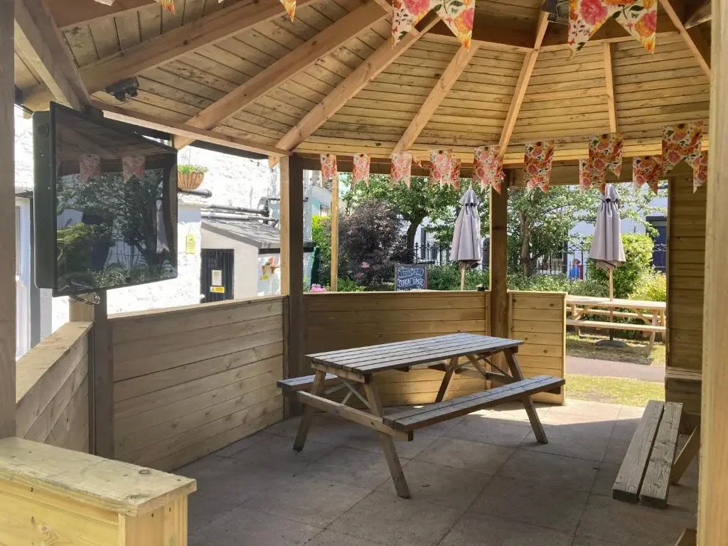 An outdoor seating area with a wooden gazebo, a picnic table, two umbrellas, a standing TV, and decorative bunting.