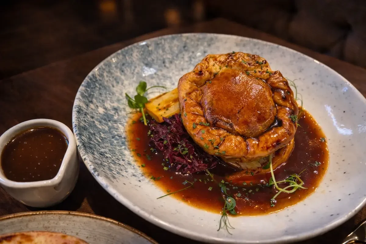 A plated gourmet dish featuring a piece of meat topped with sauce, served with vegetables and sauce on a speckled white plate, with a small pitcher of gravy on the side.