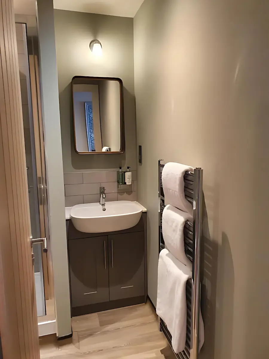 A small bathroom with a wall-mounted mirror, a sink with dark cabinet below, a lit wall sconce, and a towel rack with white towels. There is a wooden door on the left and a vertical towel heater on the right.