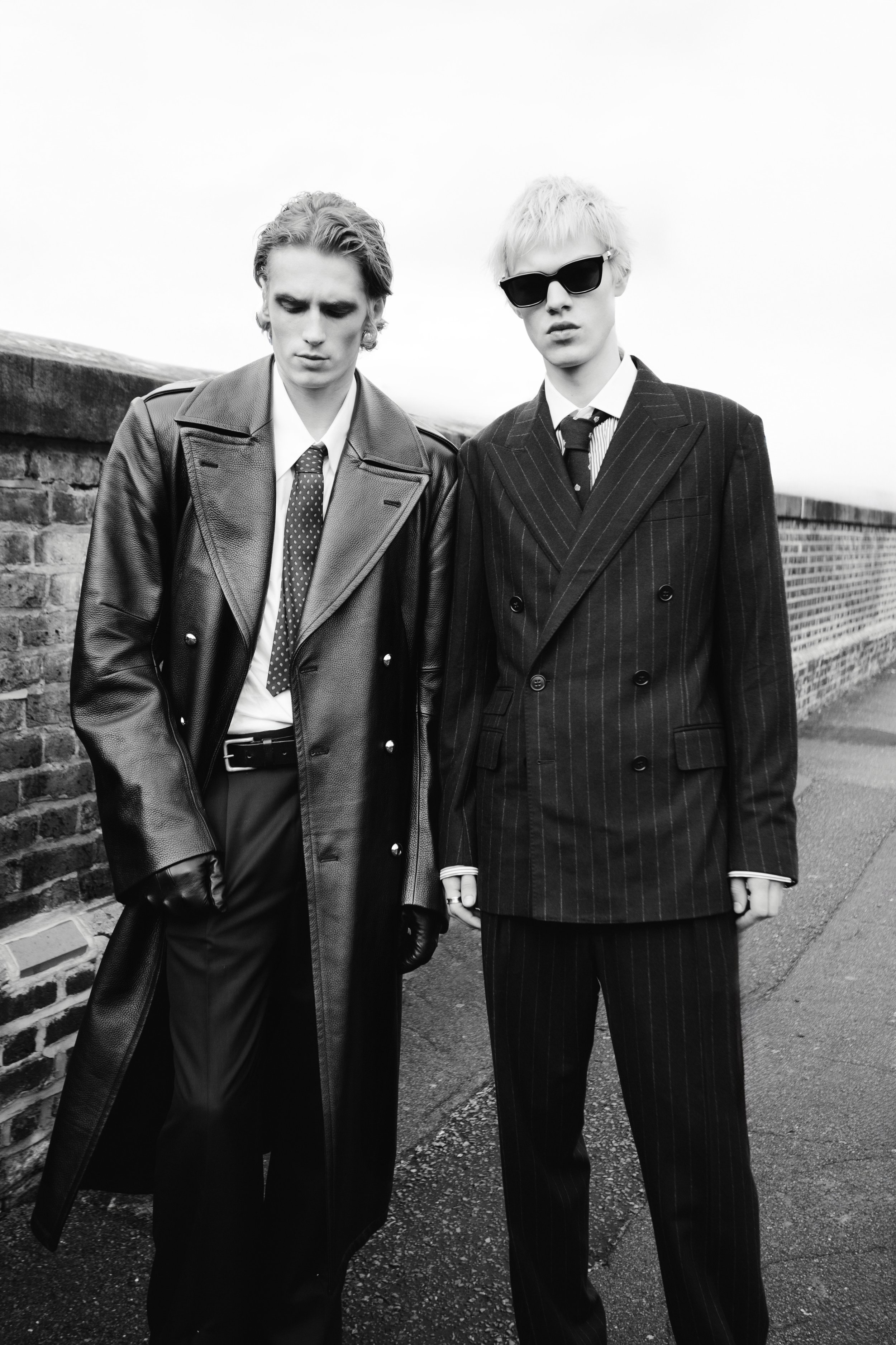Two young men in vintage black and white fashion with sunglasses, standing outdoors in front of a brick and stone wall.
