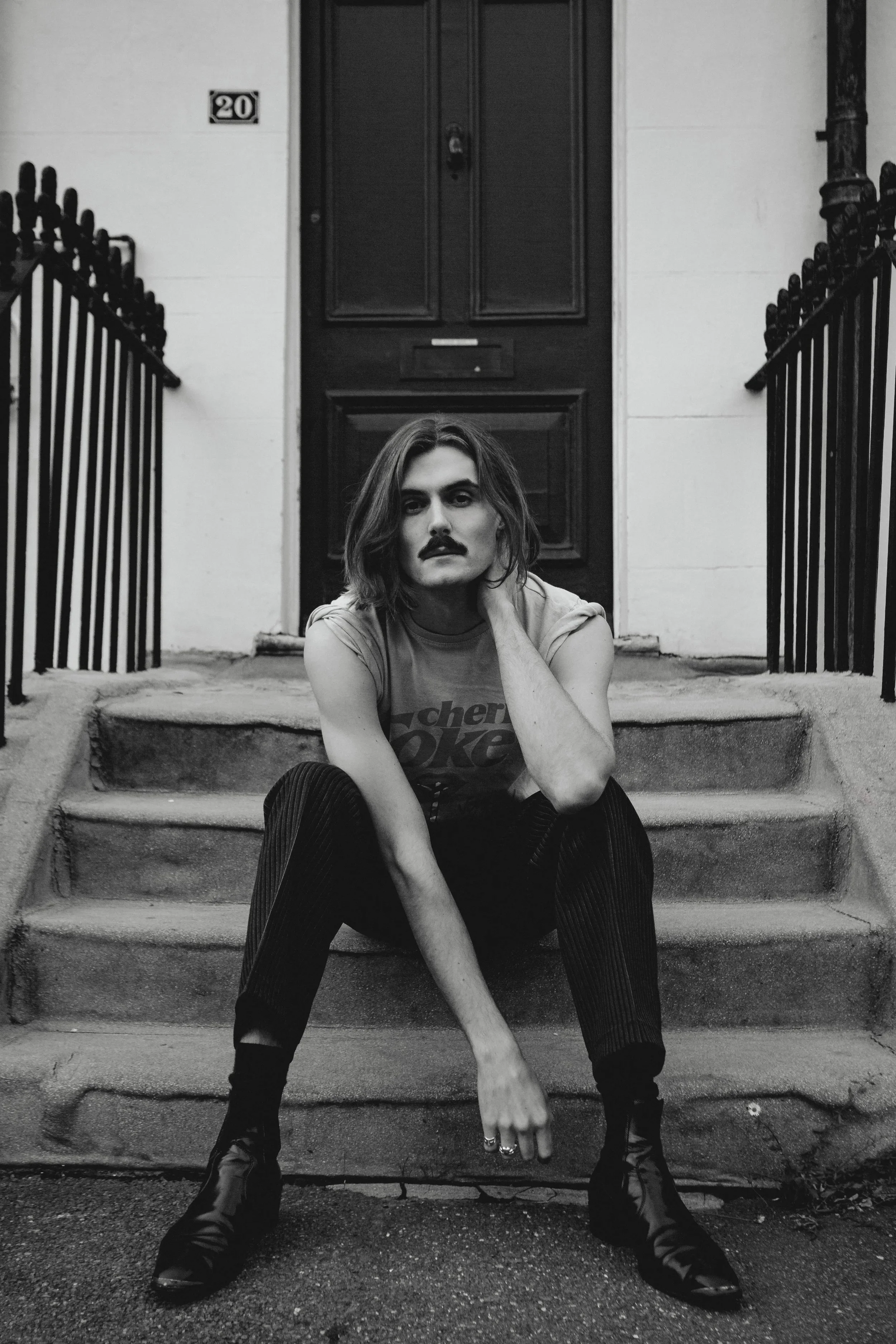 A person with shoulder-length hair, a mustache, and a confident expression, sitting on outdoor concrete steps in front of a dark door with a house number 20, flanked by metal railings, wearing a t-shirt, pants, and boots.