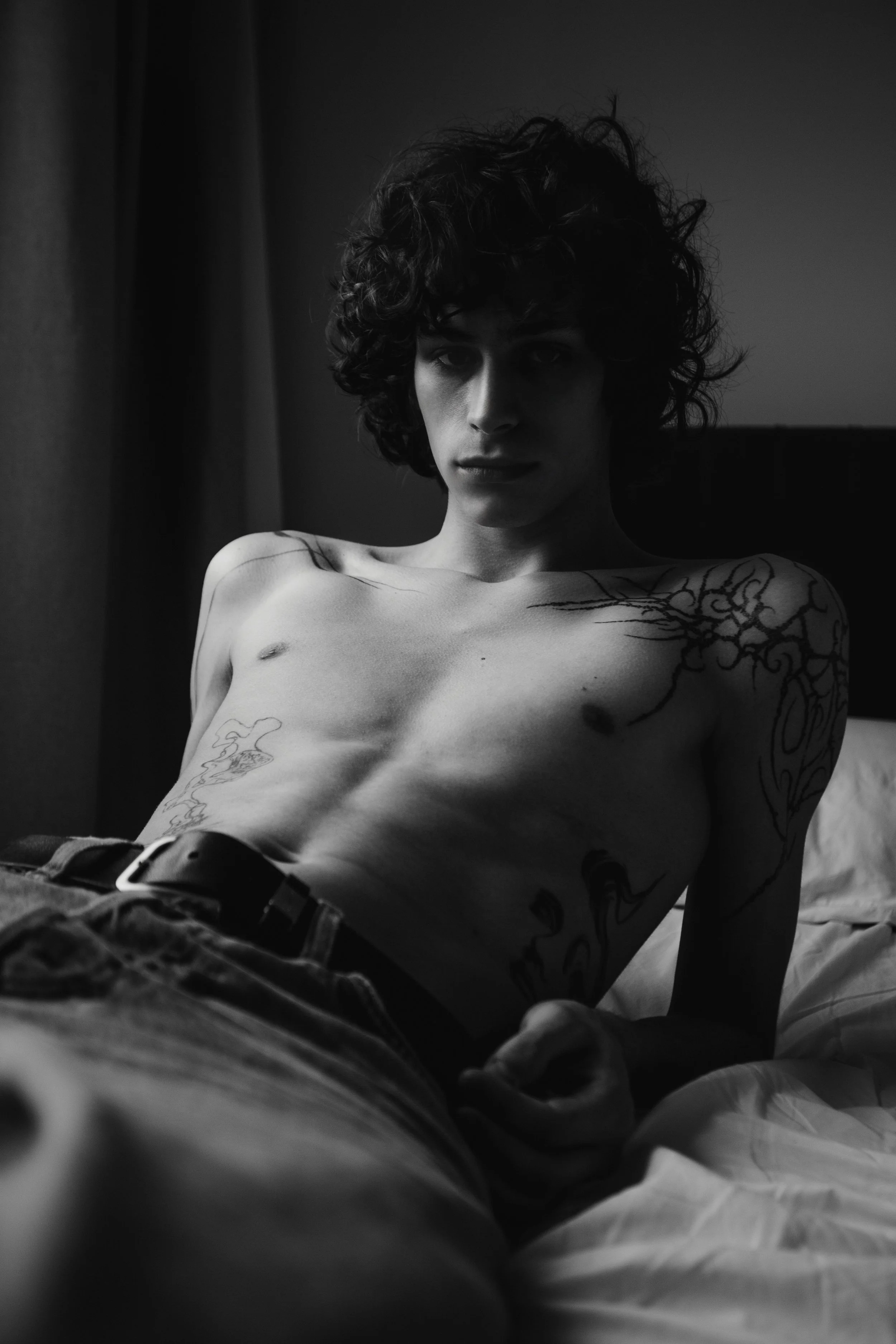 A black-and-white photo of a shirtless person with curly hair, lying on a bed, looking directly at the camera, with tattooed arm and chest.