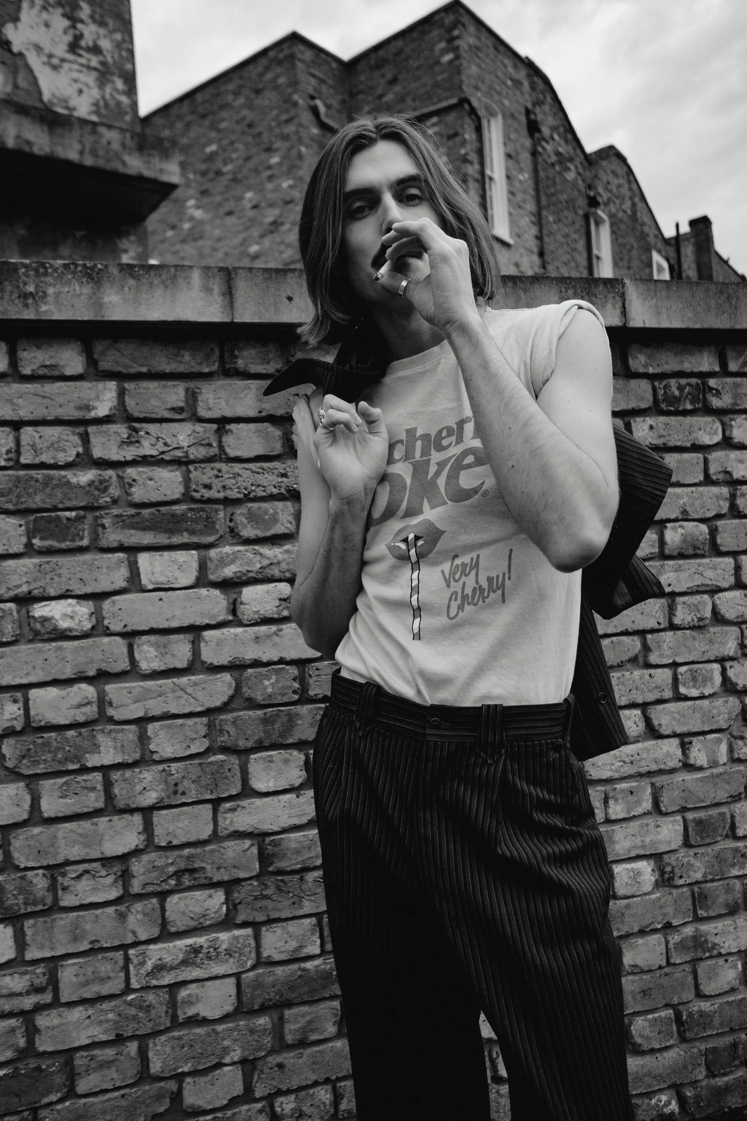 A person with shoulder-length hair is standing outdoors in front of a brick wall, holding a backpack and a cigarette, wearing a graphic t-shirt and striped pants, in black and white.
