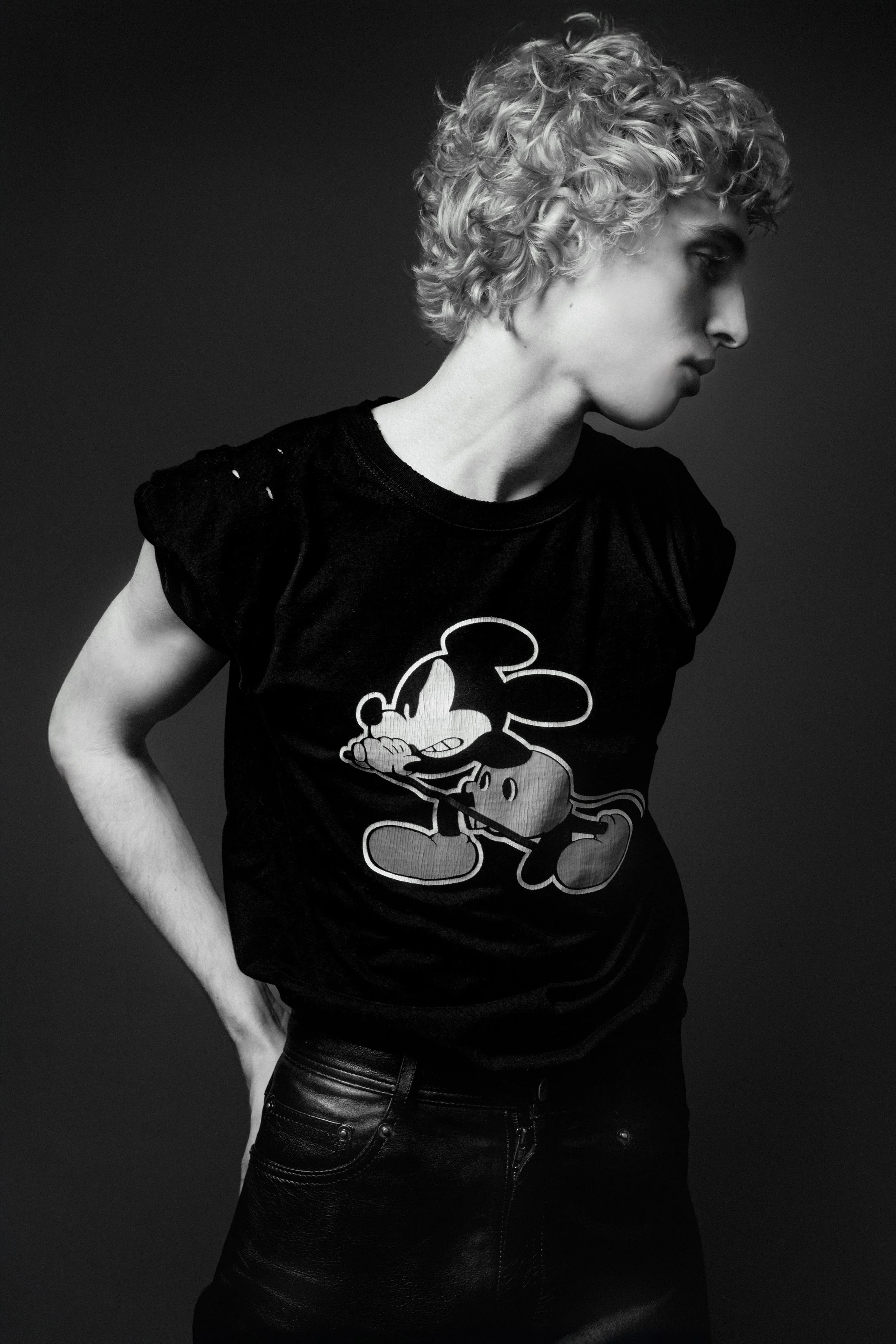 Black and white photo of a person with curly hair wearing a Mickey Mouse t-shirt and leather pants, standing against a dark background.