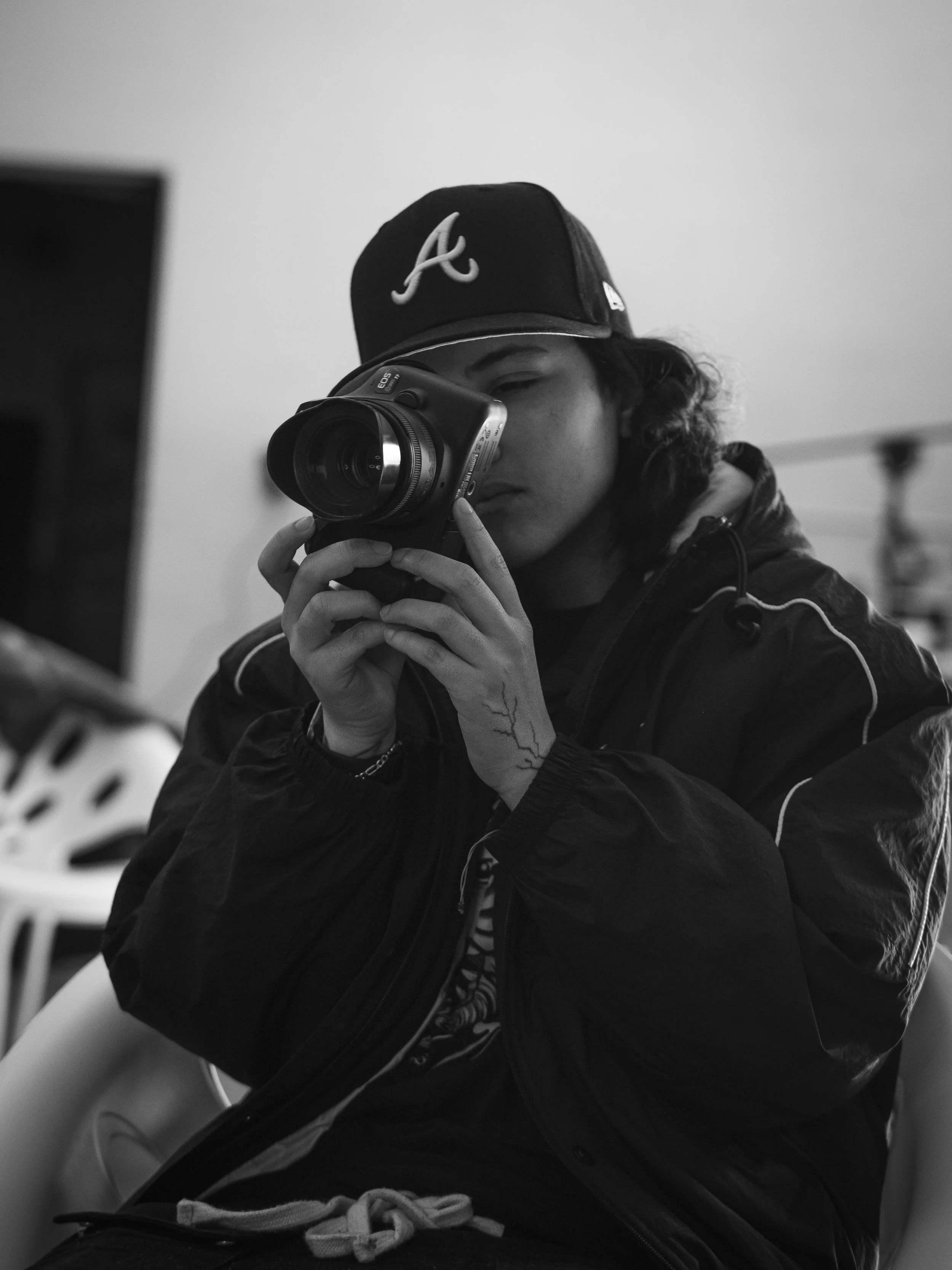 Black and white photo of a young man taking a picture with a camera, wearing a cap with an 'A' logo and a black jacket.