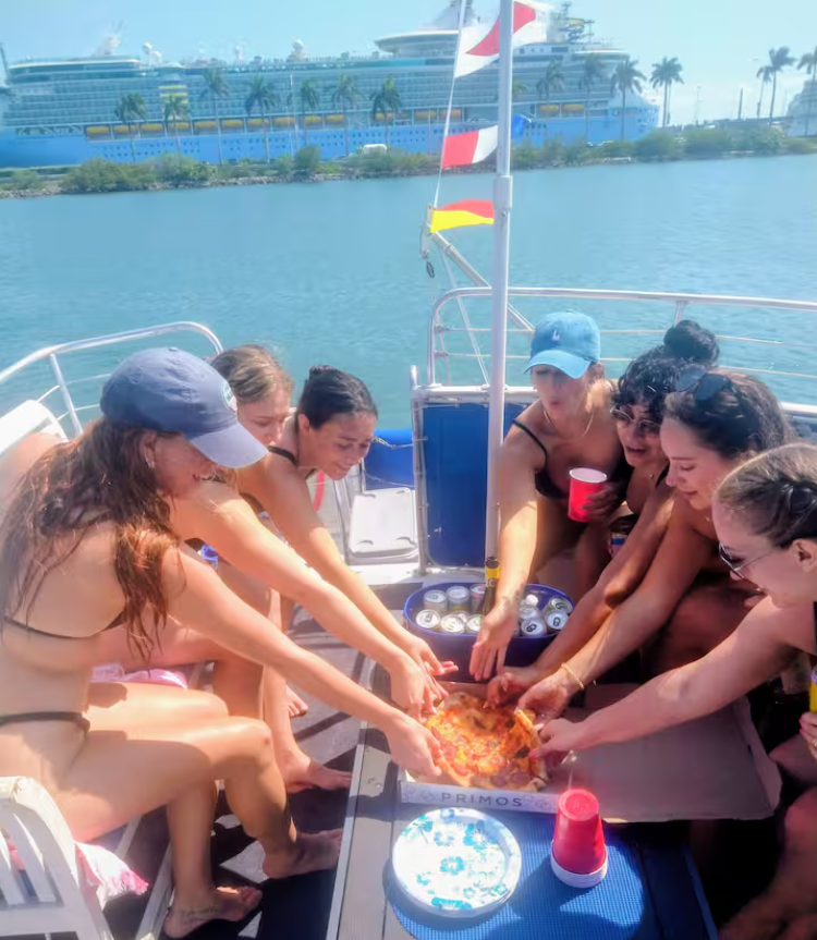 Group of young women in swimsuits and hats sharing a slice of pizza on a boat, with a cruise ship and palm trees in the background.