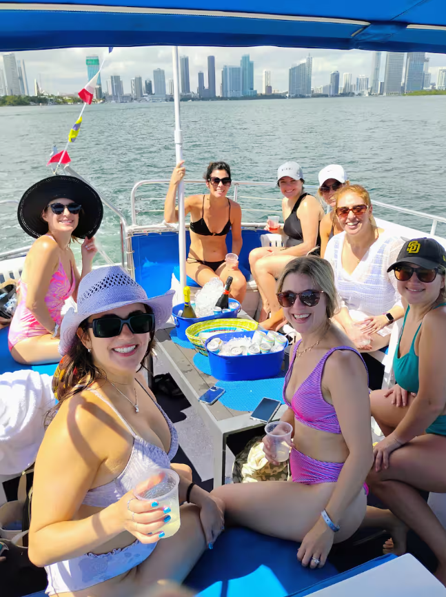 A group of nine women in swimsuits and summer hats enjoying a boat ride on a river, with a city skyline in the background.