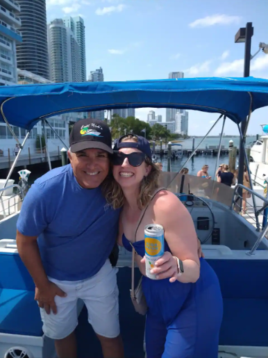 A man and woman smiling and posing on a boat with a city skyline in the background. The woman is holding a canned beverage and wearing sunglasses, and both are dressed casually.
