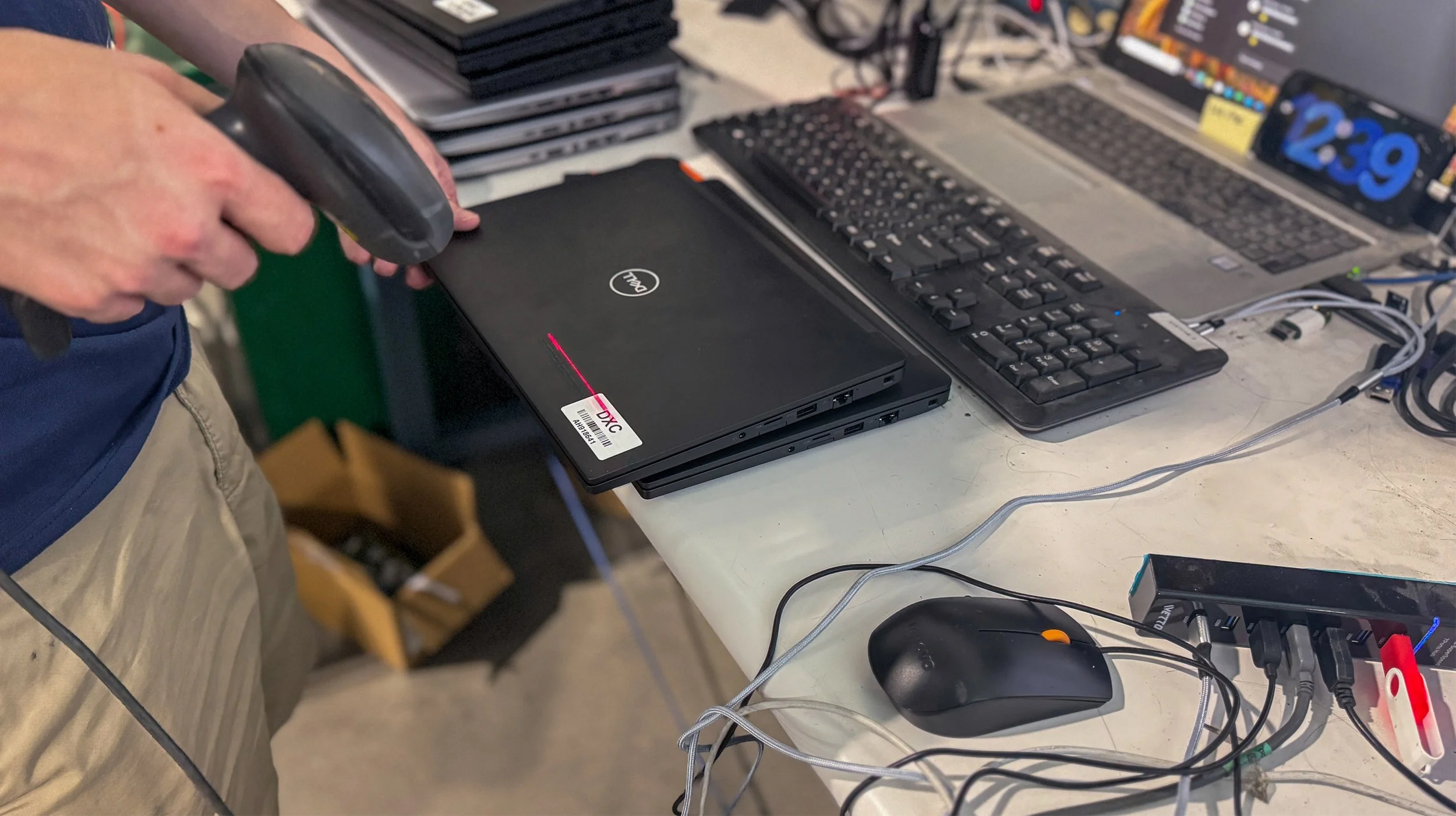 Person holding a barcode scanner over a closed Dell laptop on a cluttered desk with keyboards, a mouse, cords, and other electronic equipment.