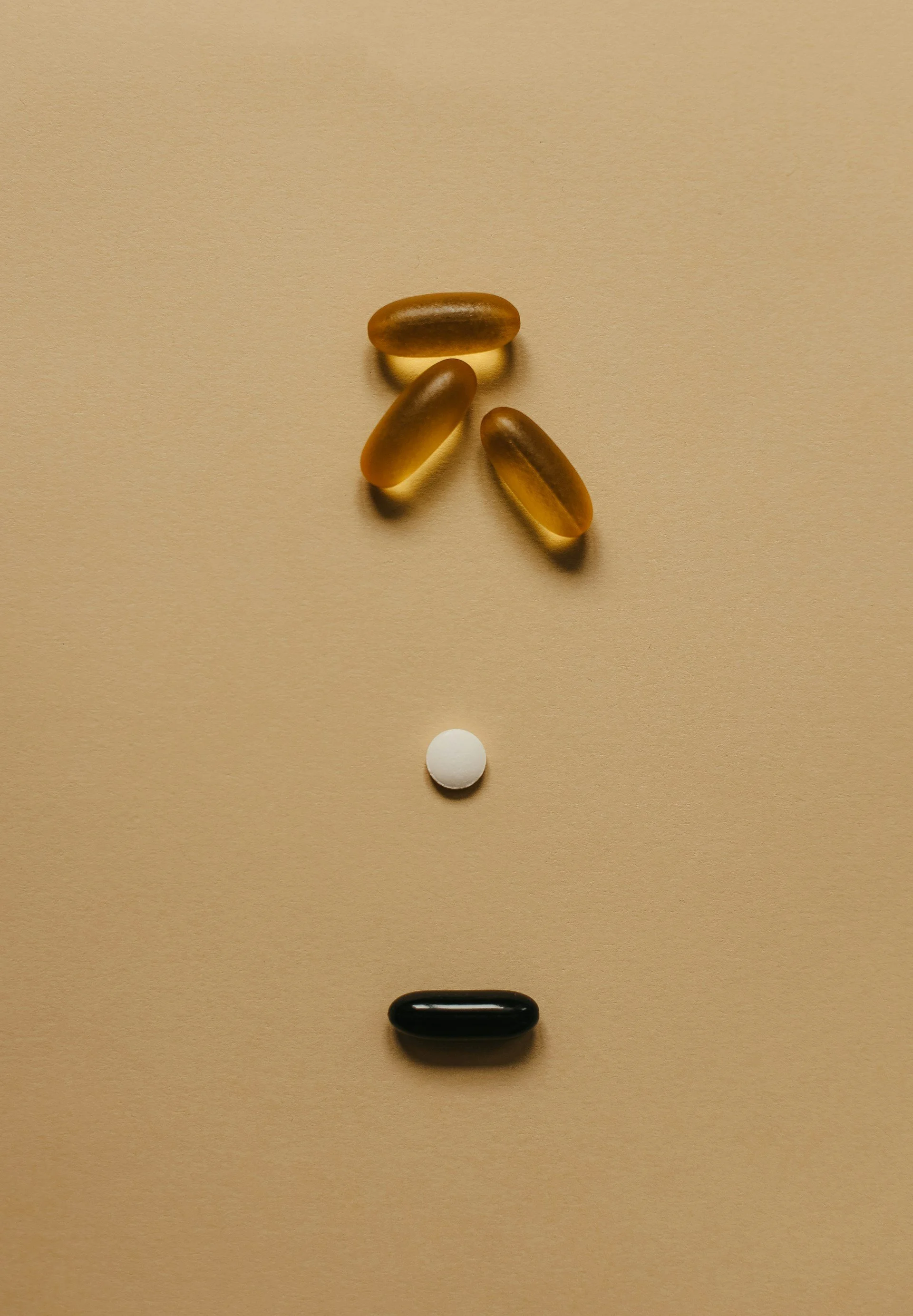 Arrangement of pills on a plain beige background, with three brown capsules at the top, a white tablet in the middle, and a black capsule at the bottom, forming a face-like design.