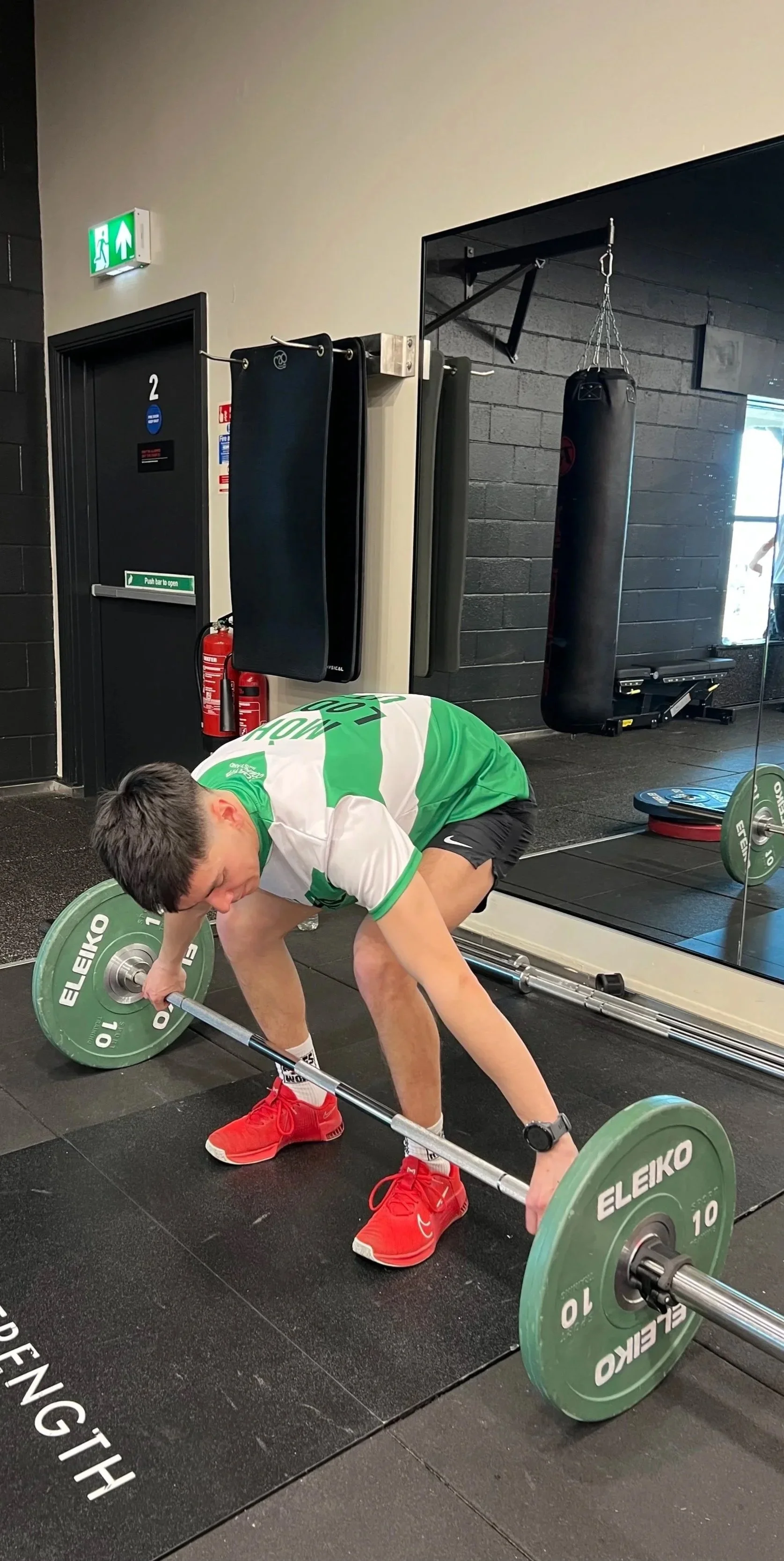 A man is lifting a barbell with green weight plates in a gym, bending over with his knees slightly bent, wearing a green and white shirt, black shorts, red shoes, and a watch.