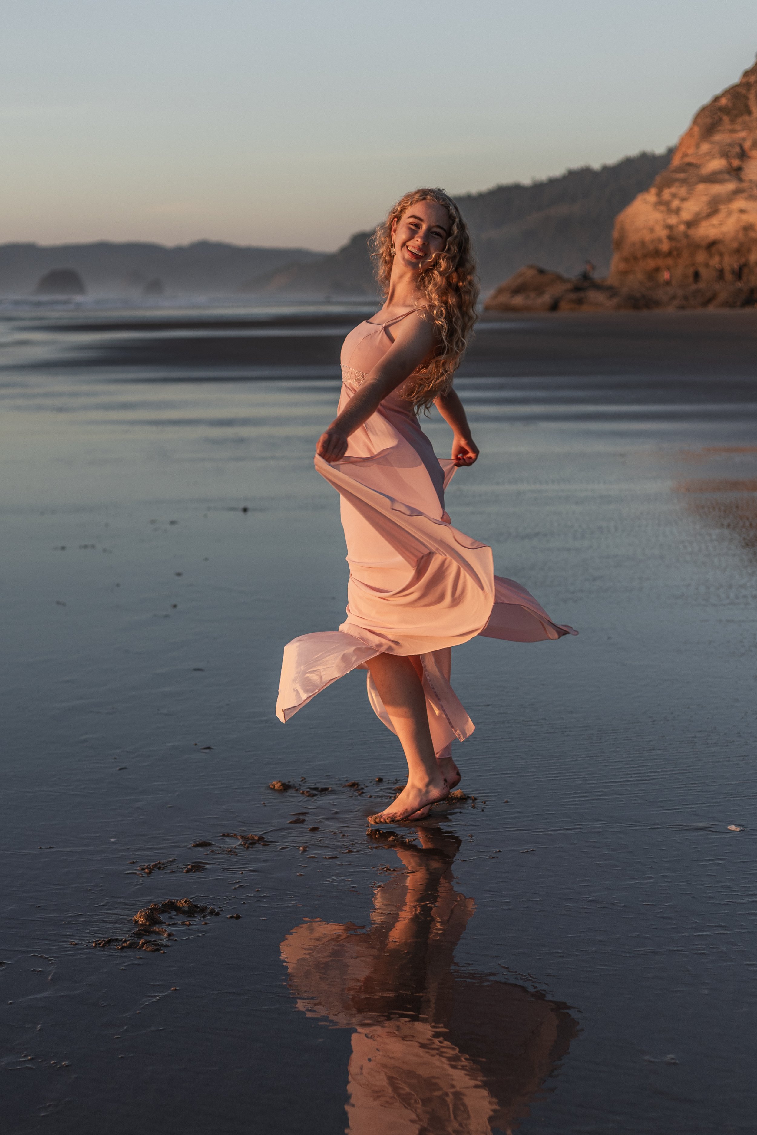 A woman in a pink dress dancing barefoot on a beach at sunset, with cliffs and ocean waves in the background.