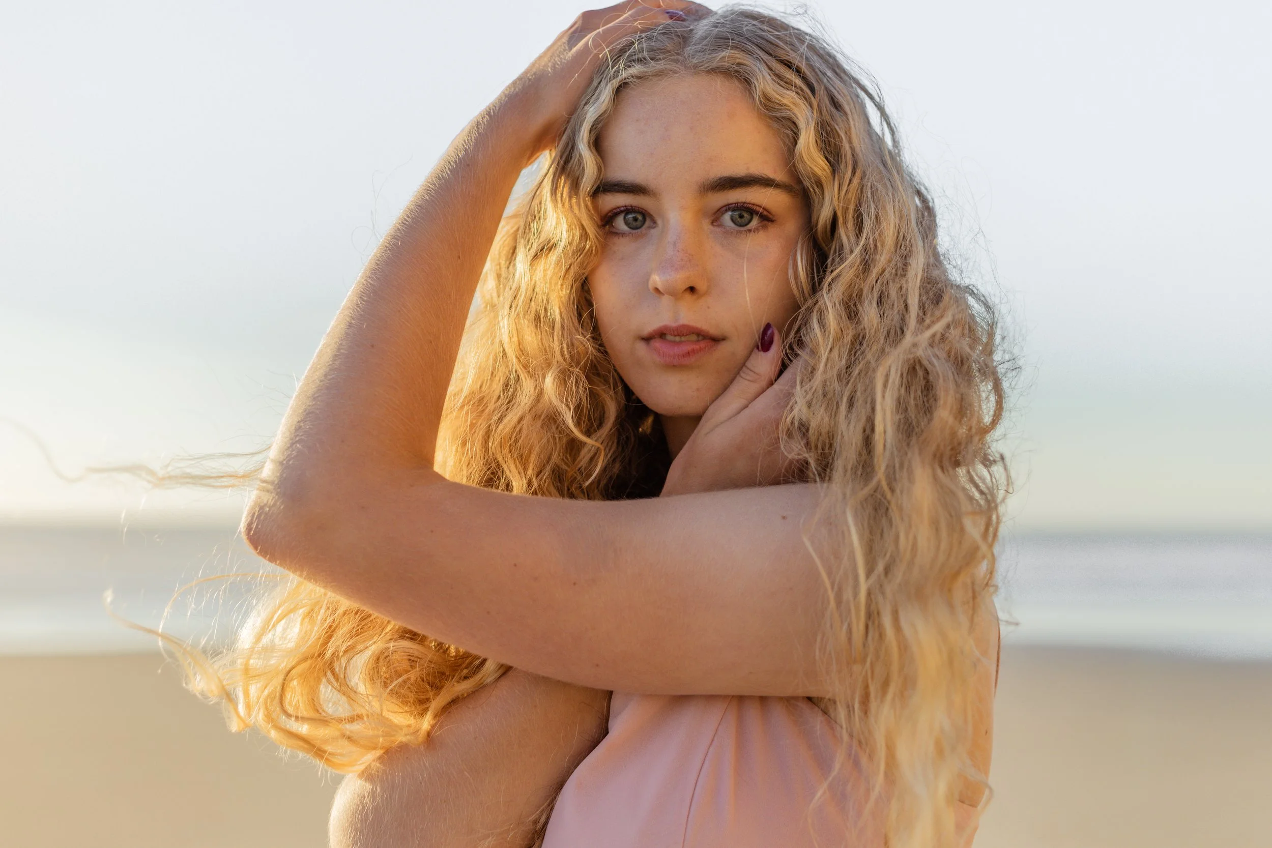 A young woman with long, curly blonde hair and blue eyes posing on the beach during sunset.