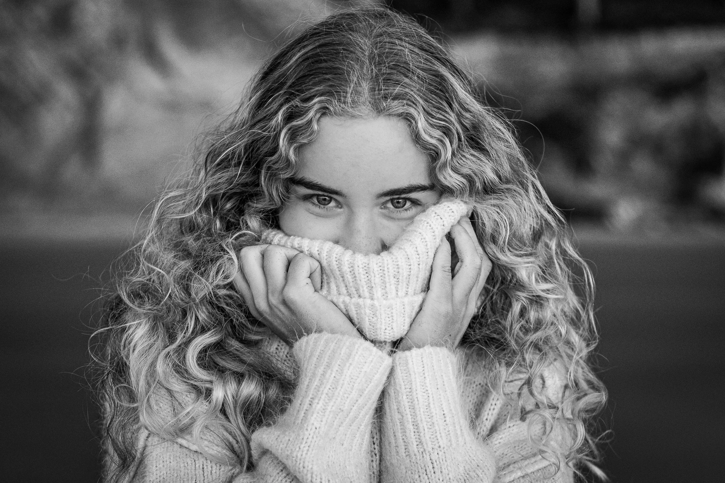 Black and white close-up portrait of a young woman with curly hair, partially covering her face with a knit sweater, looking directly at the camera.