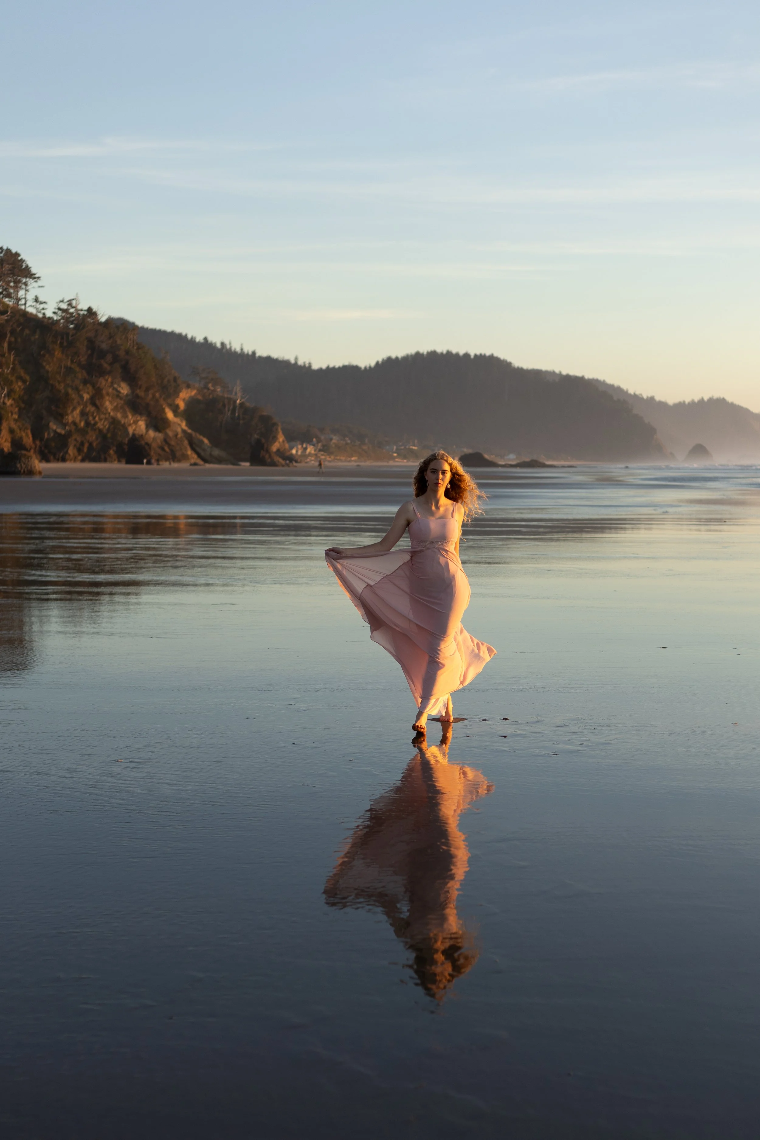 A woman in a flowing pink dress standing on wet sand, with her reflection visible in the shallow water, near the beach at sunset with hills and distant rocks in the background.